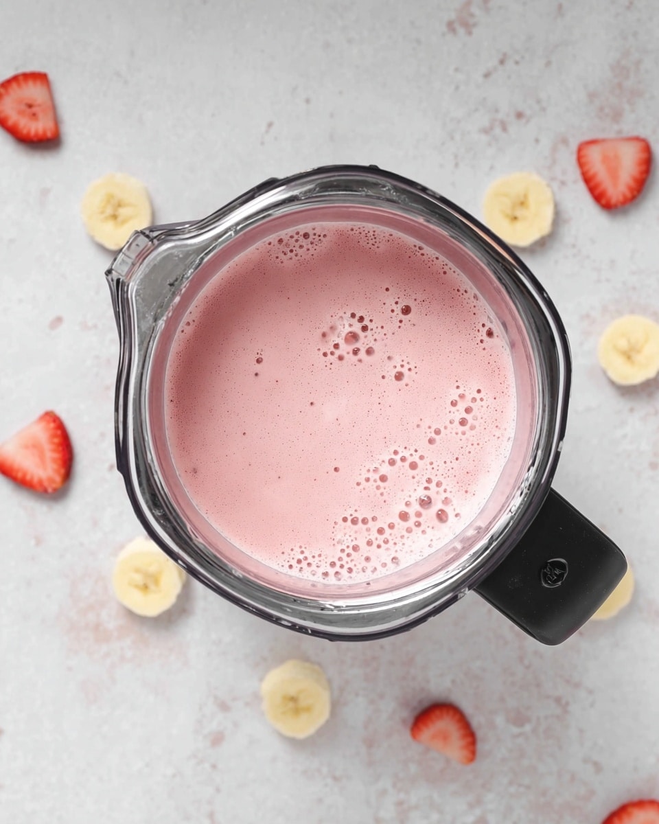 A top-down view of a blender jug filled with a smooth, pink liquid that has small bubbles on the surface, indicating a fresh smoothie mix. The jug has a black handle on the right side and shiny metal edges around the top. Around the blender, on a white marbled surface, are scattered slices of banana and pieces of strawberry, adding color contrast with yellow and red tones against the white background. The scene is simple and clean, focusing on the fresh ingredients and the blended smoothie inside the jug. photo taken with an iphone --ar 4:5 --v 7