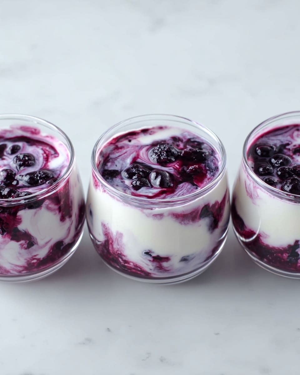 Three small clear glass bowls are placed in a line on a white marbled surface. Each bowl contains a dessert with two main layers swirled together: a smooth white creamy layer and a dark purple berry sauce with visible whole berries, creating a marbled pattern throughout. The texture looks soft and creamy with some chunks from the berries mixed in. photo taken with an iphone --ar 4:5 --v 7
