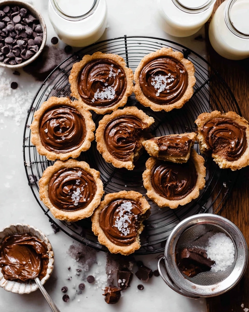 The image shows a group of small tarts arranged on a black wire cooling rack over a brown wooden surface. Each tart has a golden-brown crust filled with smooth, glossy chocolate filling that has a swirled texture on top. One tart in the center is cut in half, showing a soft, dense chocolate center. Scattered around the tarts are small dark chocolate chips, a white bowl of coarse salt, and a metal sieve filled with more chocolate chips. There are three glass bottles of milk placed around the tarts and a silver spoon with some chocolate spread nearby. The scene is set on a white marbled surface. photo taken with an iphone --ar 4:5 --v 7