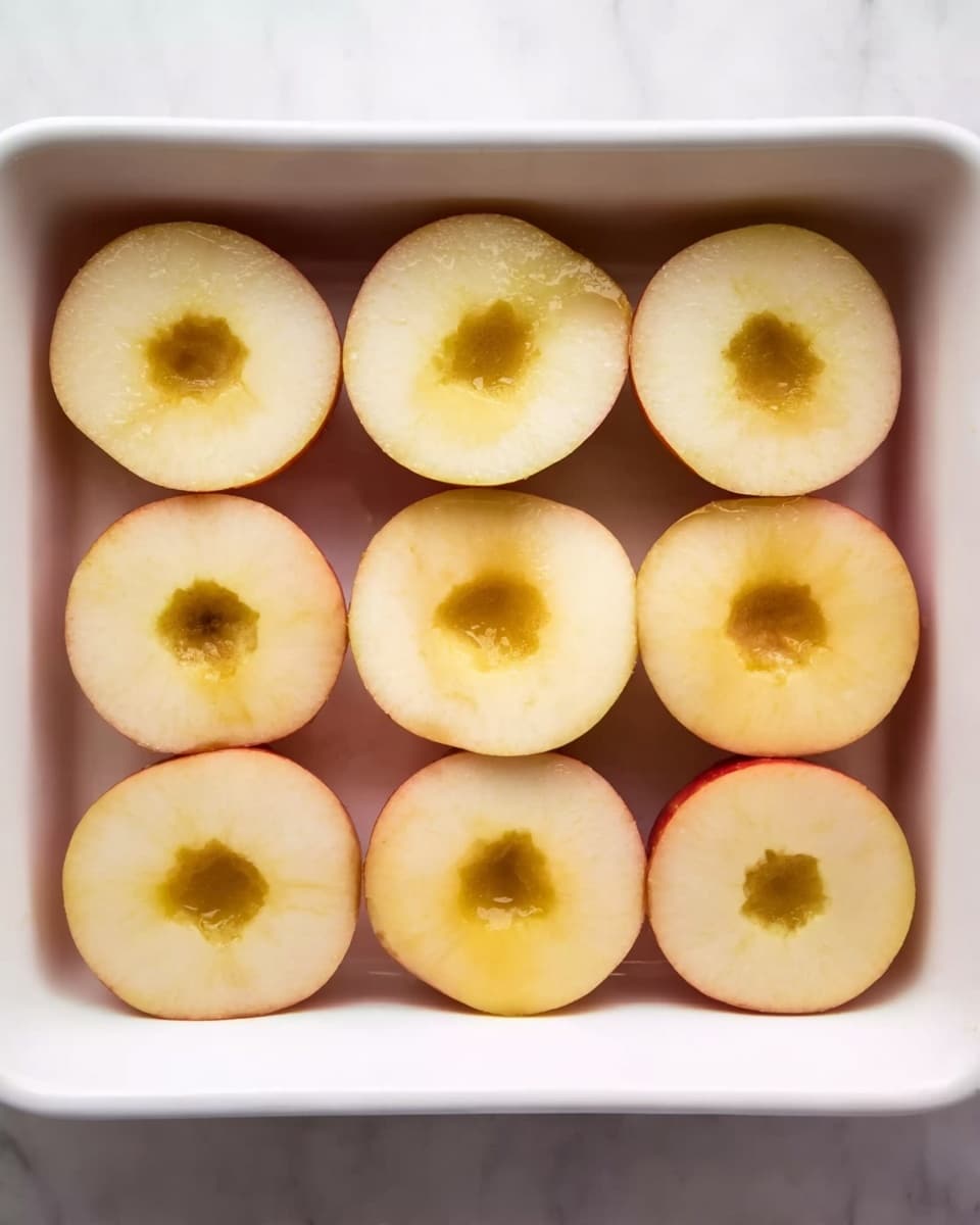 The image shows a white baking dish filled with two layers of apple halves arranged in neat rows. The apple halves have their cores removed, creating hollow circles in the center, with the skin showing a pale yellow color on the inside and a light reddish outer edge. The apples rest directly on the white dish, and some of the hollow centers have a slight golden brown shade. The background is a white marbled texture. Photo taken with an iphone --ar 4:5 --v 7