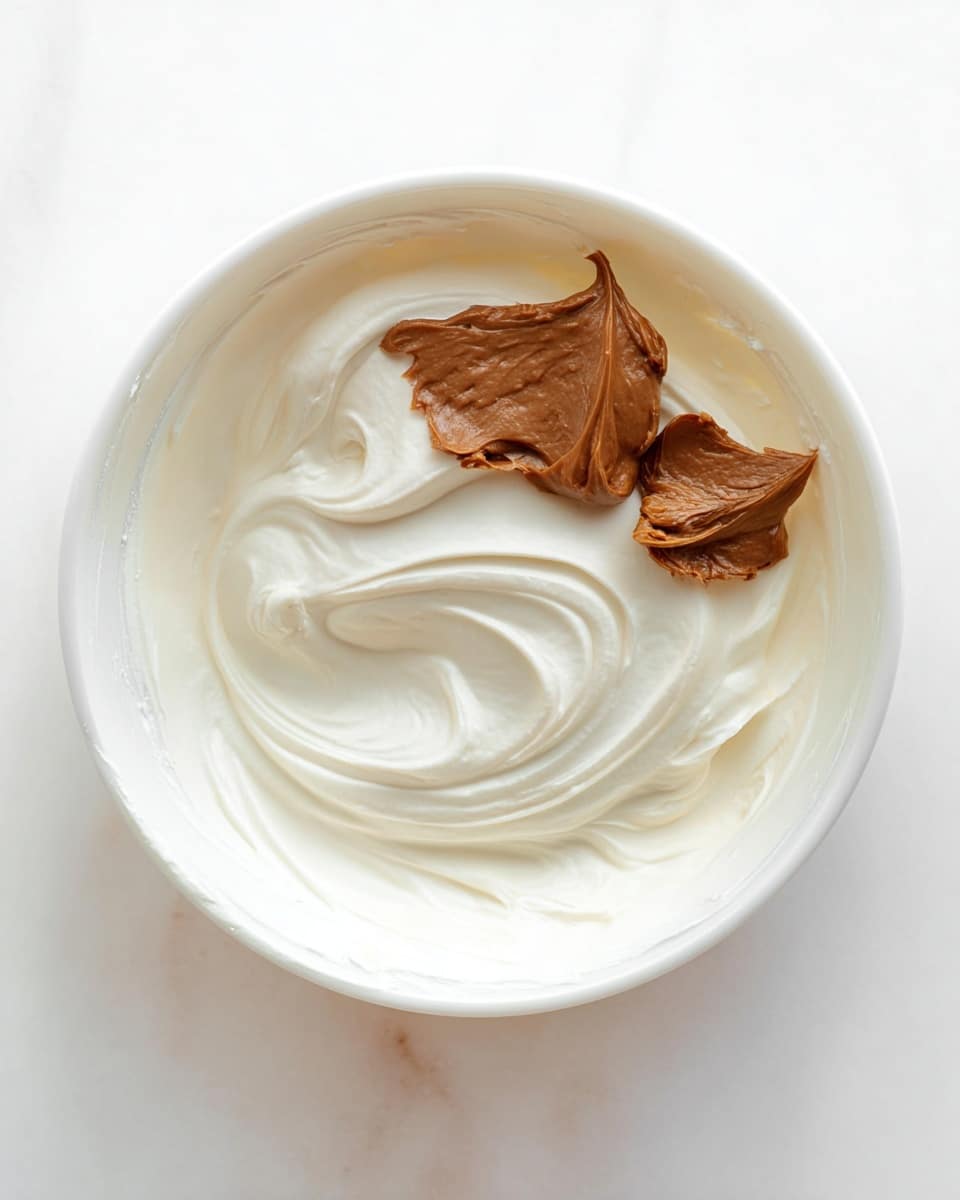 A white bowl filled with a thick, smooth white cream forming the base layer, with swirls creating a soft texture around the inside edges. On top, there are two dollops of brown paste, one larger and smoother and the other smaller and slightly textured, placed close to the center right. The bowl sits on a white marbled surface. Photo taken with an iphone --ar 4:5 --v 7