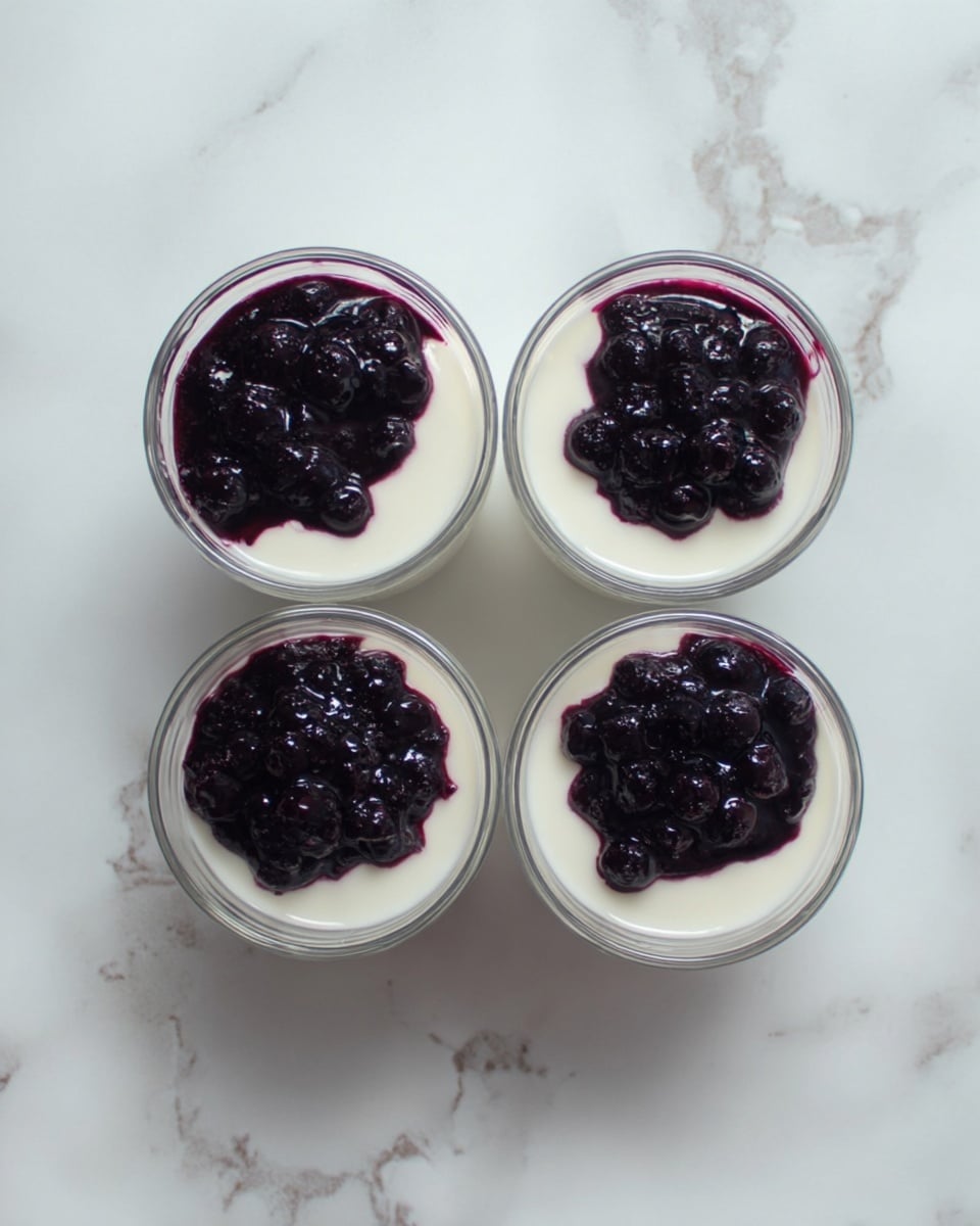 Four small clear glass cups are placed on a white marbled surface. Each cup has two layers: a bottom layer of smooth white yogurt and a top layer of dark purple blueberry sauce with small whole blueberries visible. The blueberry layer covers roughly half of the yogurt surface in each cup. The cups are arranged in a square, with two at the top and two at the bottom. photo taken with an iphone --ar 4:5 --v 7