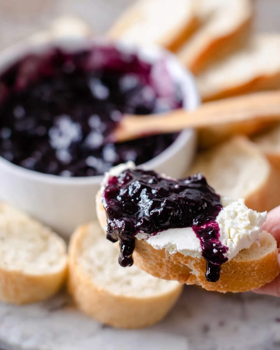 The image shows a small piece of light golden bread topped with thick white cream cheese and a dark purple-blue jelly spread, slightly dripping on the side, held by a woman's hand. Behind it, there is a white round dish filled with the same dark purple-blue jelly with a wooden spreader resting on the edge. In the background, there are several slices of light-colored bread arranged loosely on a white marbled surface. The colors of the bread, cream cheese, and jelly create a simple but appealing contrast. photo taken with an iphone --ar 4:5 --v 7