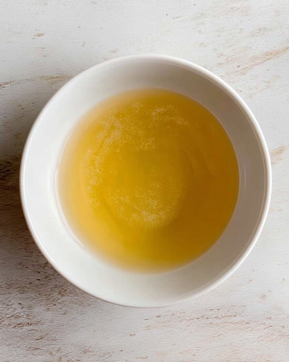 A white bowl holds a clear golden-yellow liquid with a slightly cloudy texture, sitting on a white marbled surface. The liquid fills most of the bowl, with a smooth surface showing subtle light reflections. The bowl is centered and viewed from above, showing only its round rim and the shine of the liquid inside. Photo taken with an iphone --ar 4:5 --v 7