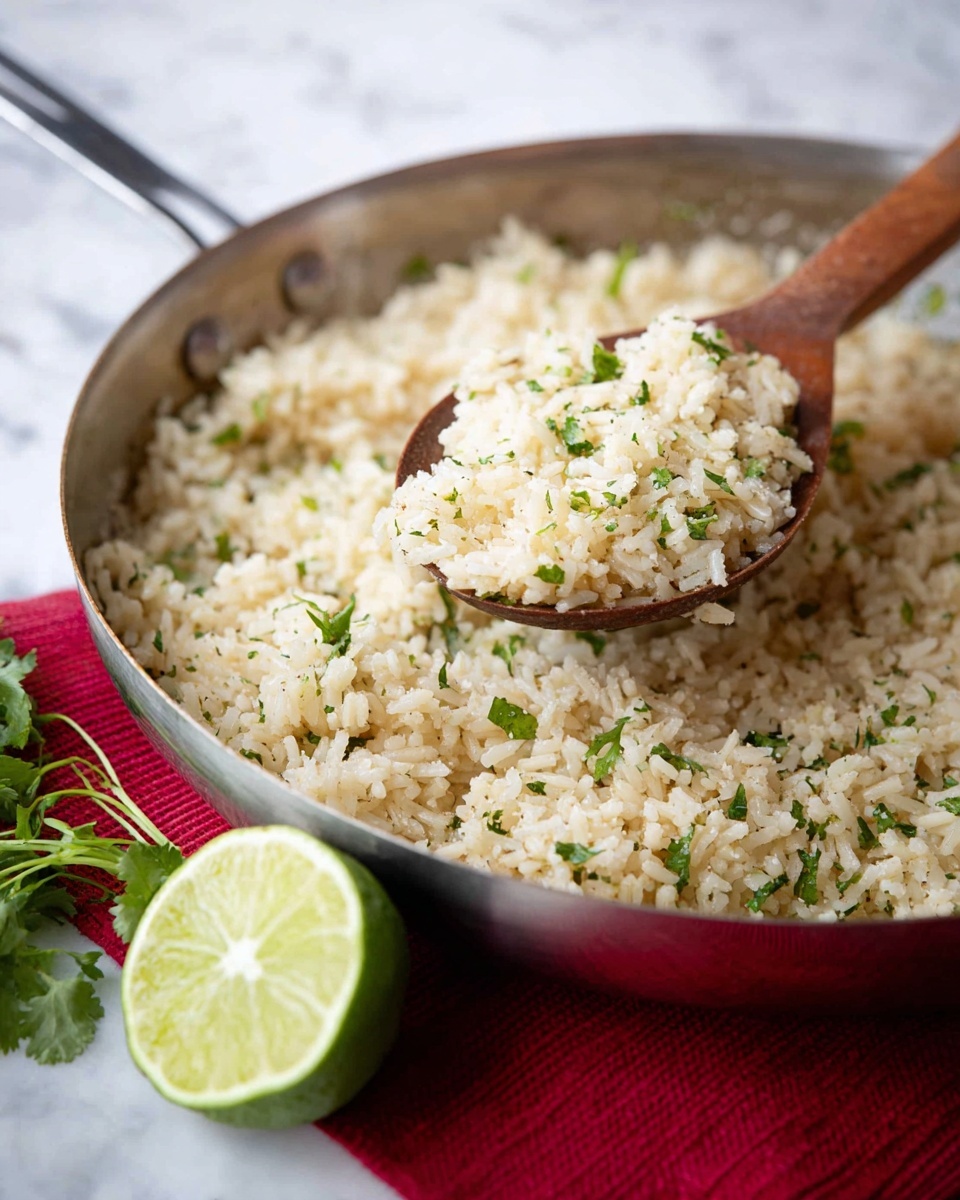 A close-up view of a metal pan filled with a single layer of light beige cooked rice mixed with small green herb pieces scattered throughout. Above the pan, a wooden spoon holds a scoop of the same rice, showing its grainy and soft texture with green herb bits. In front of the pan, a halved lime with a light green interior sits on a white marbled surface. Fresh green herbs rest near the lime and pan, with a red cloth faintly visible under the pan. Photo taken with an iphone --ar 4:5 --v 7