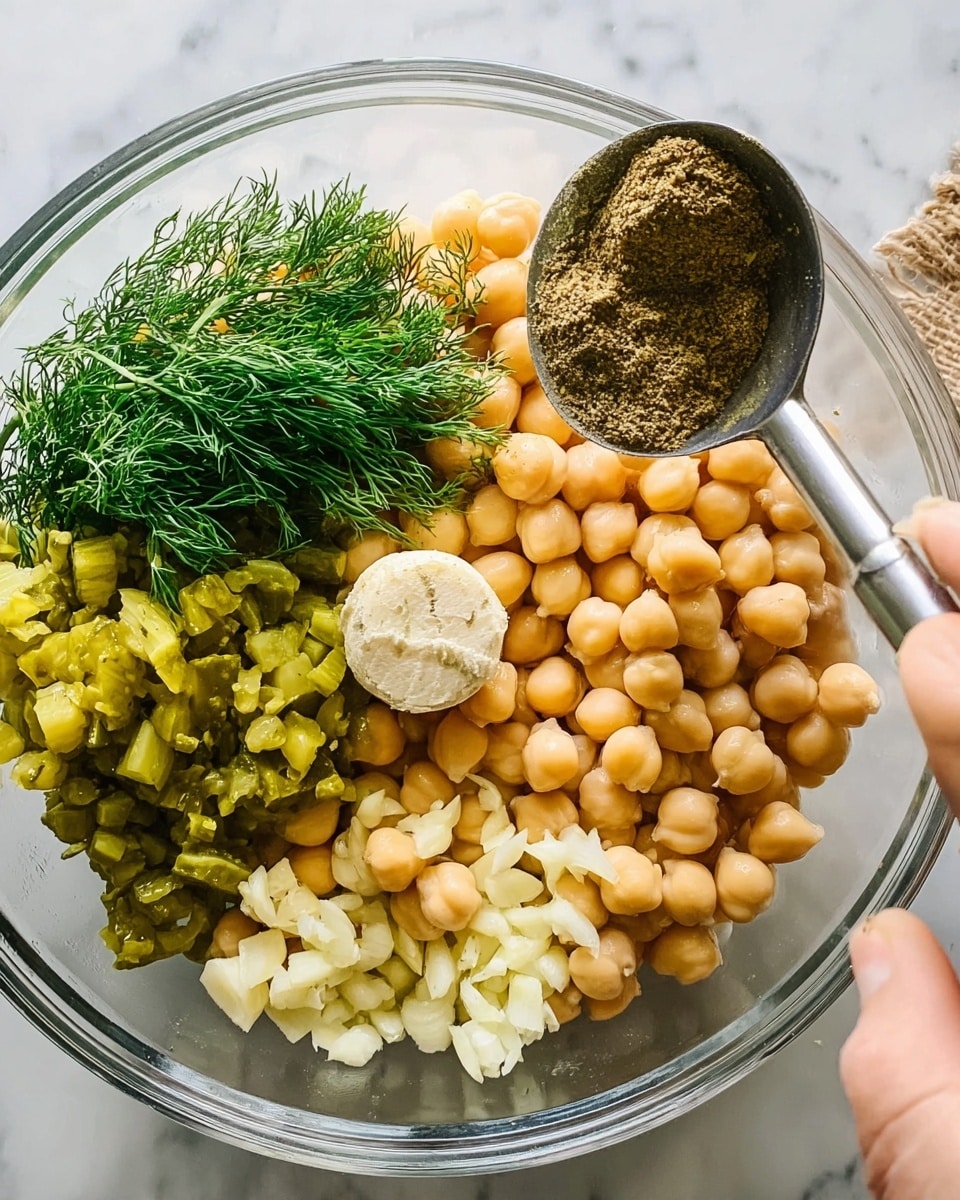 A clear glass bowl is filled mostly with light beige chickpeas, which cover most of the bottom and right side. On the left bottom is a pile of chopped bright green pickles, near the top left are fresh, bright green dill sprigs stacked together, and in the center rests a small round piece of smooth off-white garlic paste. Near the bottom right of the chickpeas is a small pile of chopped white garlic pieces. Above the bowl on the right side, a woman's hand holds a silver measuring spoon with a scoop of dark greenish-brown spice. The background and surface are white marbled texture. photo taken with an iphone --ar 4:5 --v 7