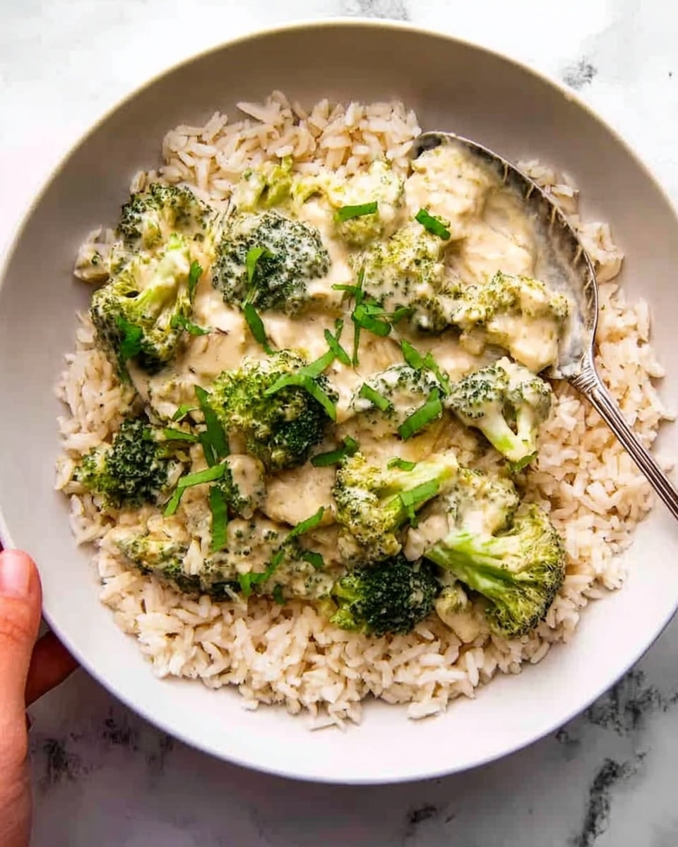 A white bowl holds a layer of light brown cooked rice at the bottom, topped with a creamy white sauce mixed with bright green broccoli florets and chunks of light-colored chicken. Small sprigs of fresh green herbs are scattered on top, adding a touch of brightness. A silver spoon rests on the right side of the bowl, partially covered by the food. Behind the bowl, there is another white bowl filled with the same creamy broccoli and chicken mix. The scene is set on a white marbled surface, with a textured gray and green striped cloth on the left side and a small glass jar with green herbs in the upper left corner. photo taken with an iphone --ar 4:5 --v 7
