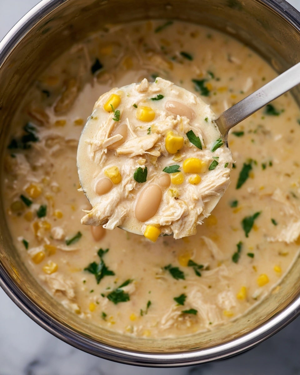 The image shows a close-up of a creamy soup in a shiny stainless steel pot. The soup is thick and light beige in color, filled with pieces of shredded chicken, white beans, and yellow corn kernels. Small green herbs are scattered on top, adding a fresh touch of color. A large silver ladle lifts a portion of the soup, showing the mix of ingredients clearly, with the creamy texture holding them together. The background is a white marbled texture, emphasizing the warm and hearty look of the soup. photo taken with an iphone --ar 4:5 --v 7