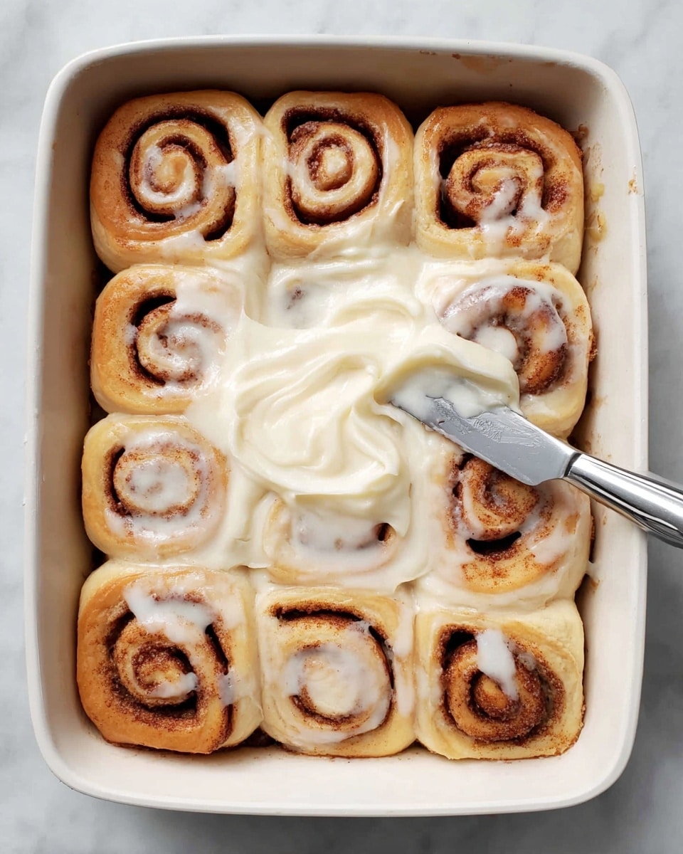 The image shows a white baking dish filled with twelve cinnamon rolls arranged in three rows of four, all touching each other. Each roll has a soft, light beige dough outer layer with a dark brown cinnamon swirl inside, creating tight spiral patterns visible from the top. The rolls look soft and unbaked, with smooth dough and slightly uneven shapes giving a homemade feel. The dish sits on a white marbled surface, adding a clean and simple background. photo taken with an iphone --ar 4:5 --v 7