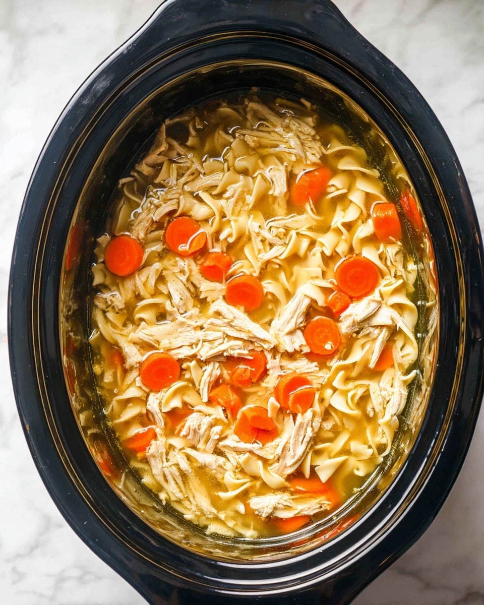 This image shows a slow cooker filled with chicken noodle soup. Inside, there are layers of light brown shredded chicken pieces evenly spread throughout the soup, with bright orange round carrot slices scattered on top and mixed in. The noodles are curved and pale yellow, sitting in a clear, golden broth that fills the cooker about halfway. The slow cooker itself is black with a shiny finish, placed on a white marbled surface. Photo taken with an iphone --ar 4:5 --v 7