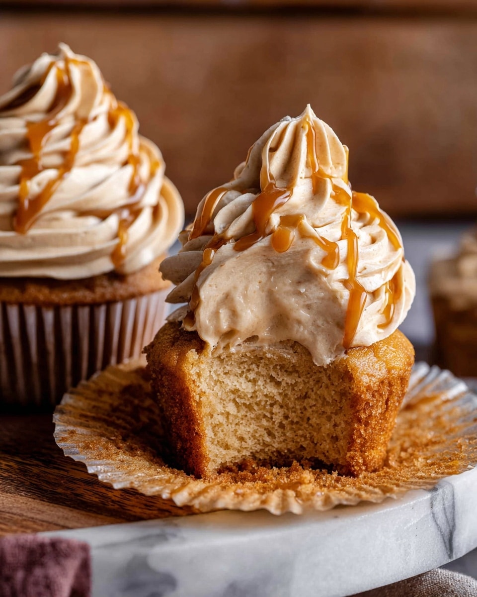 The image shows a close-up of two cupcakes on a wooden board with a soft focus background. The cupcakes have a light brown fluffy base with one cupcake partially unwrapped, revealing a moist texture. Each cupcake is topped with thick swirls of creamy beige frosting that has a smooth and rich texture. A glossy caramel drizzle is artistically spread over the frosting in thin lines, adding contrast and shine. The cupcakes are placed on a white marbled surface that complements the warm tones and rustic wood elements in the background. Photo taken with an iphone --ar 4:5 --v 7