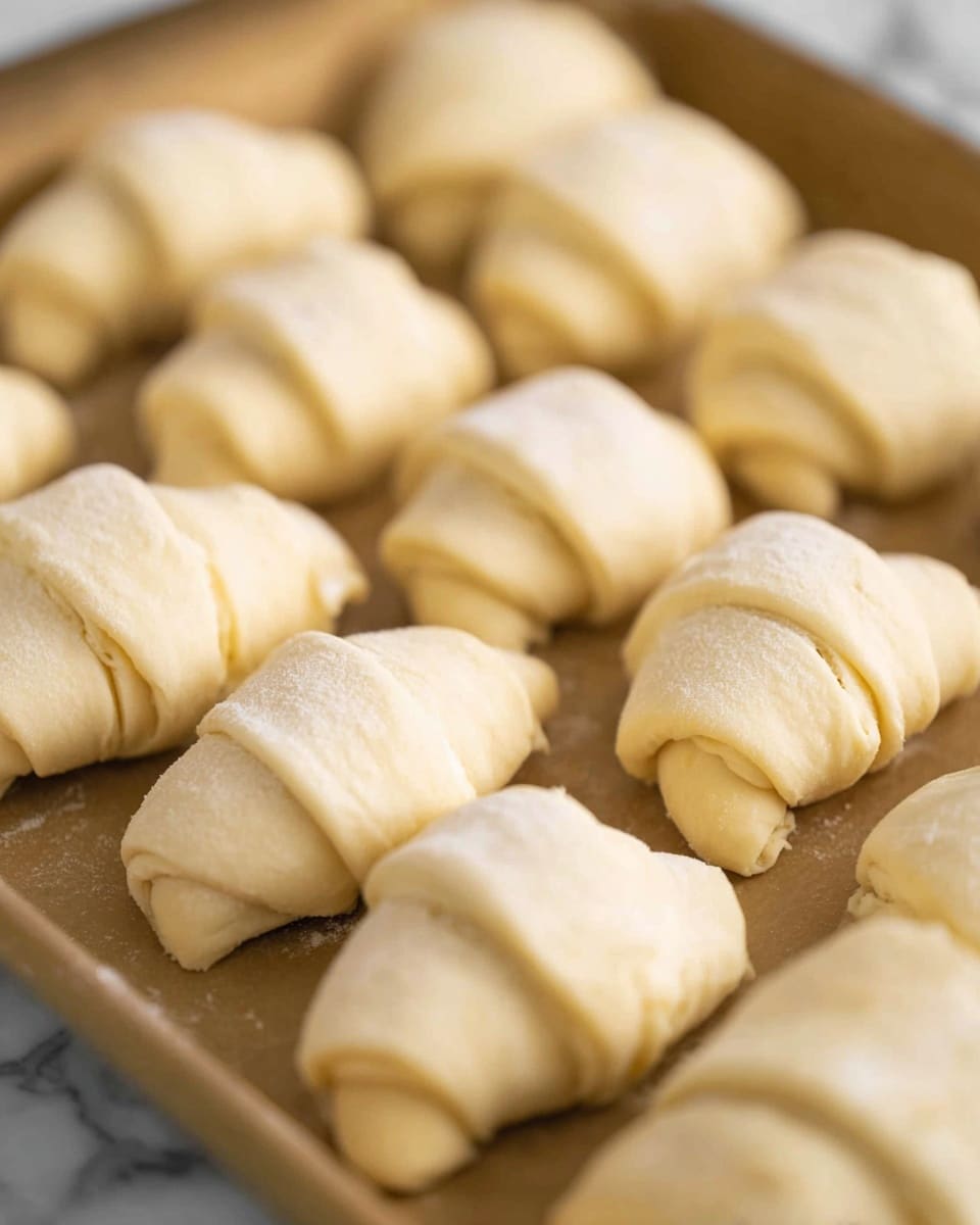 The image shows a baking tray filled with uncooked crescent rolls. Each roll is wrapped in smooth, pale dough with a soft texture, forming small, crescent shapes. The rolls are arranged in rows, placed close together but not touching, on the light brown tray. The dough looks slightly glossy in some areas with faint flour dusting, and the edges of the rolls have small folds showing where the dough has been wrapped. The background is a white marbled texture. Photo taken with an iphone --ar 4:5 --v 7