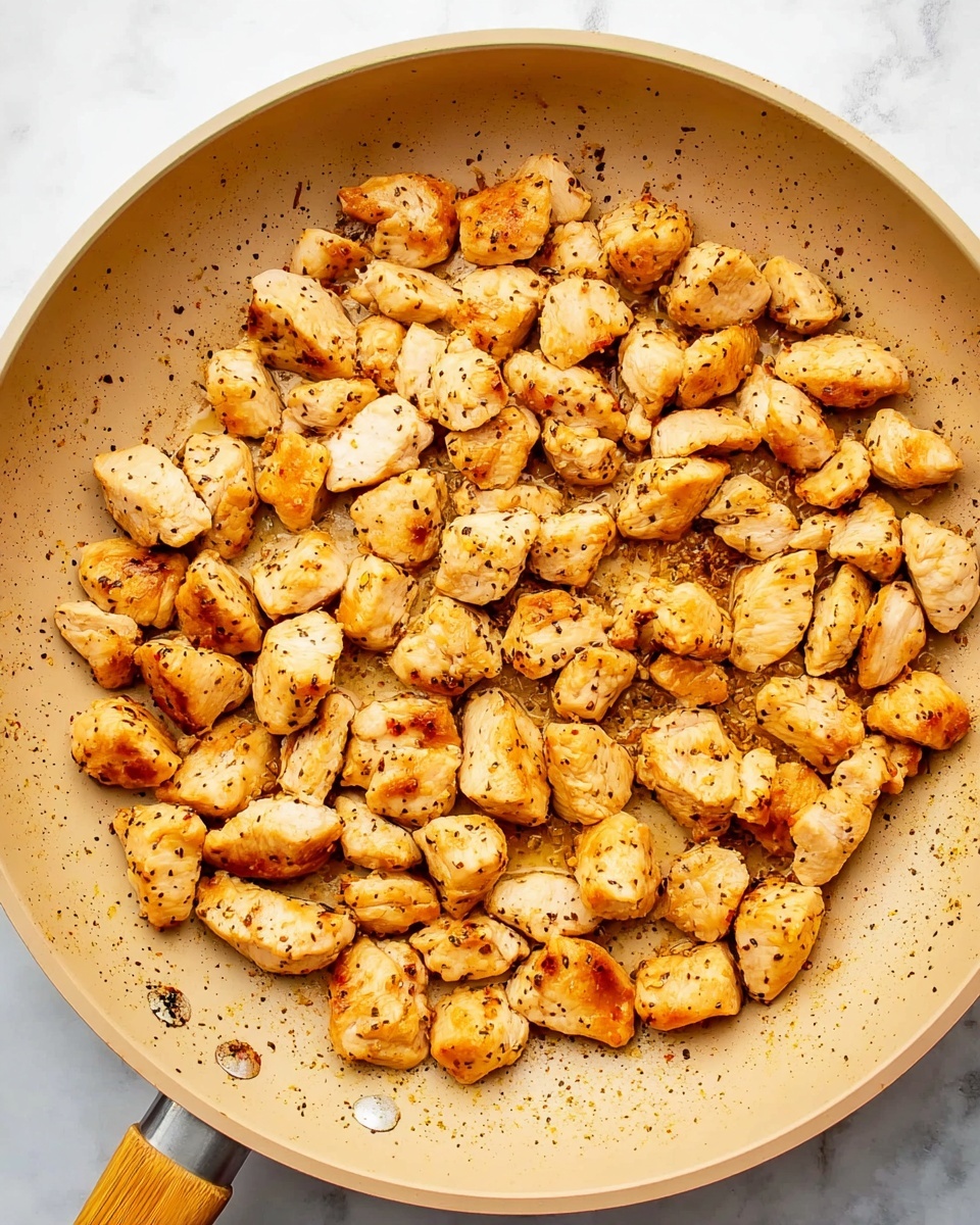 The image shows a tan frying pan filled with small pieces of cooked chicken that are light golden brown with specks of seasoning, evenly spread across the pan's surface. The pan has a light wooden handle, and the overall background is a white marbled texture. The cooked chicken pieces have a slightly crispy texture on the outside and are juicy inside, with some pieces showing more browned edges than others, creating a varied golden shade. Photo taken with an iphone --ar 4:5 --v 7