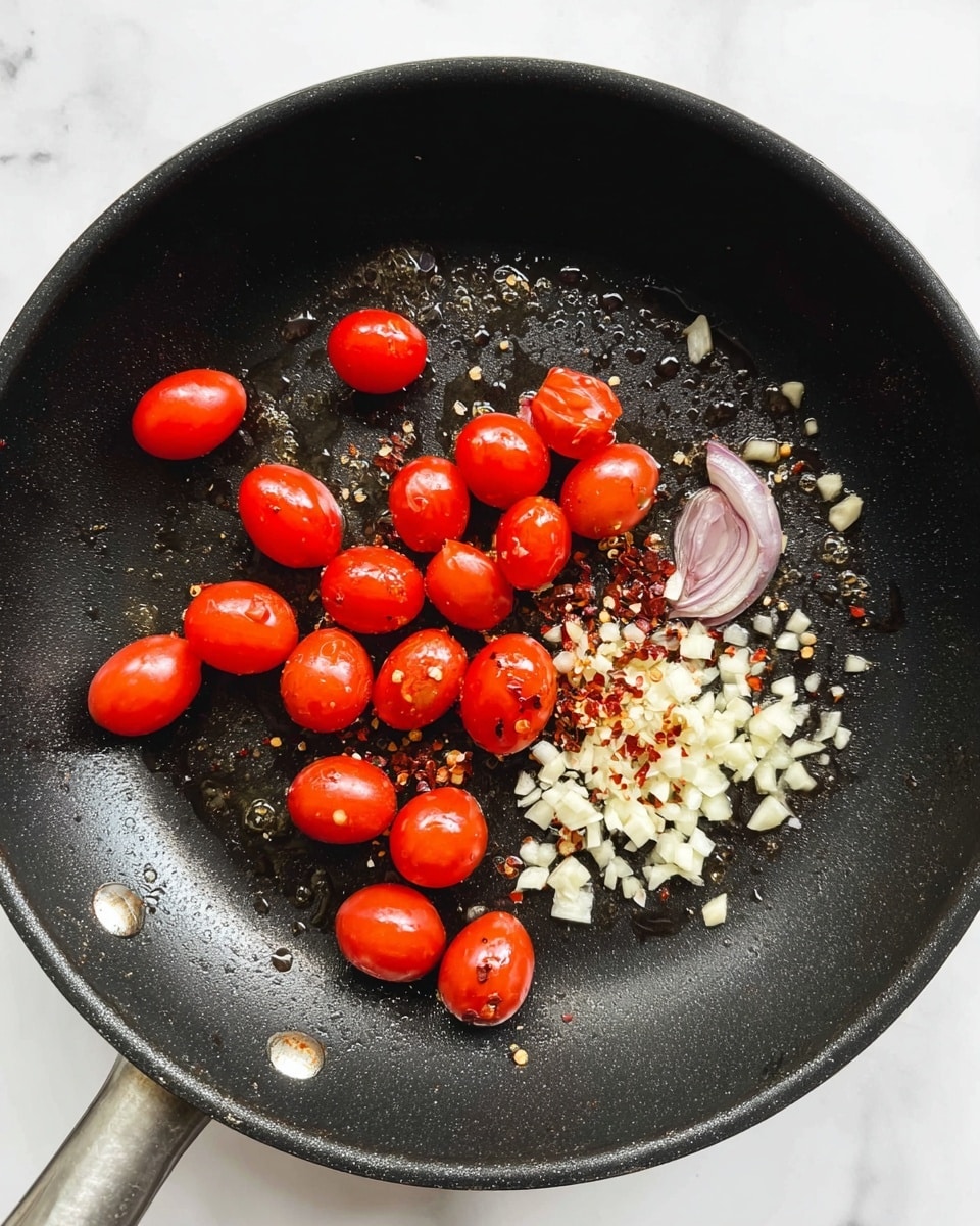 A black frying pan sits on a white marbled surface, containing three main groups of ingredients: bright red cherry tomatoes scattered across the pan, finely chopped white garlic clustered near the top center, and thin, pale purple slices of shallot near the center right. There are also small red chili flakes mixed near the garlic. The ingredients shine slightly as if coated with oil, and the pan has subtle texture marks. Photo taken with an iphone --ar 4:5 --v 7
