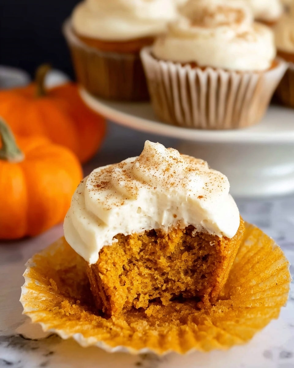 The image shows a close-up of a pumpkin cupcake with two thick layers of creamy white frosting swirled on top, sprinkled lightly with cinnamon powder. The cupcake has a light brown color with a slightly textured surface from the baking. It is wrapped in a white paper liner. Behind it, there are more cupcakes with the same frosting, placed on a white plate. The background includes a white marbled surface with a cinnamon stick and small orange pumpkins, adding a warm autumn feel. Photo taken with an iphone --ar 4:5 --v 7