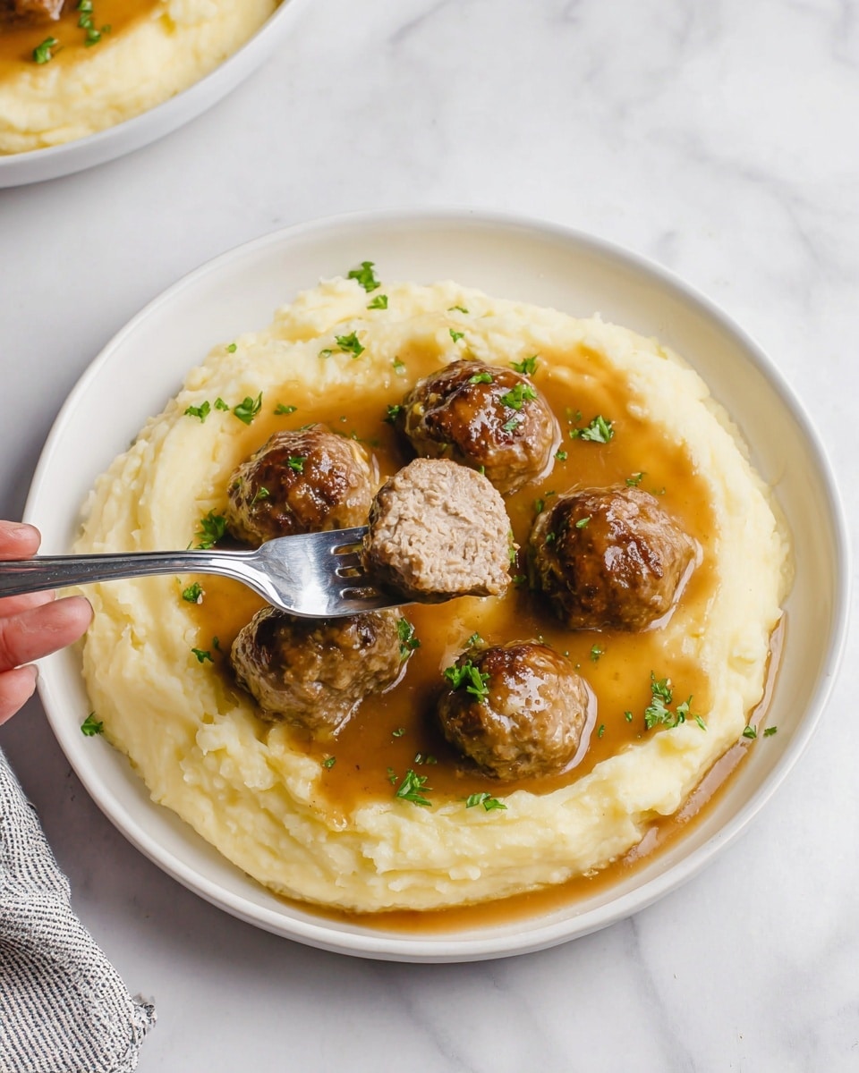 A white bowl holds a smooth, creamy layer of light yellow mashed potatoes filling the bottom and sides. On top sit three round brown meatballs with a shiny brown gravy poured over them and dripped onto the mashed potatoes. One meatball is sliced in half, showing its light brown inside with small green specks. Small green herb bits are sprinkled on top of the meatballs and potatoes. A knife with some mashed potatoes on it rests on the right side of the bowl. The bowl is set on a white marbled surface with another plate blurred in the background. Photo taken with an iphone --ar 4:5 --v 7