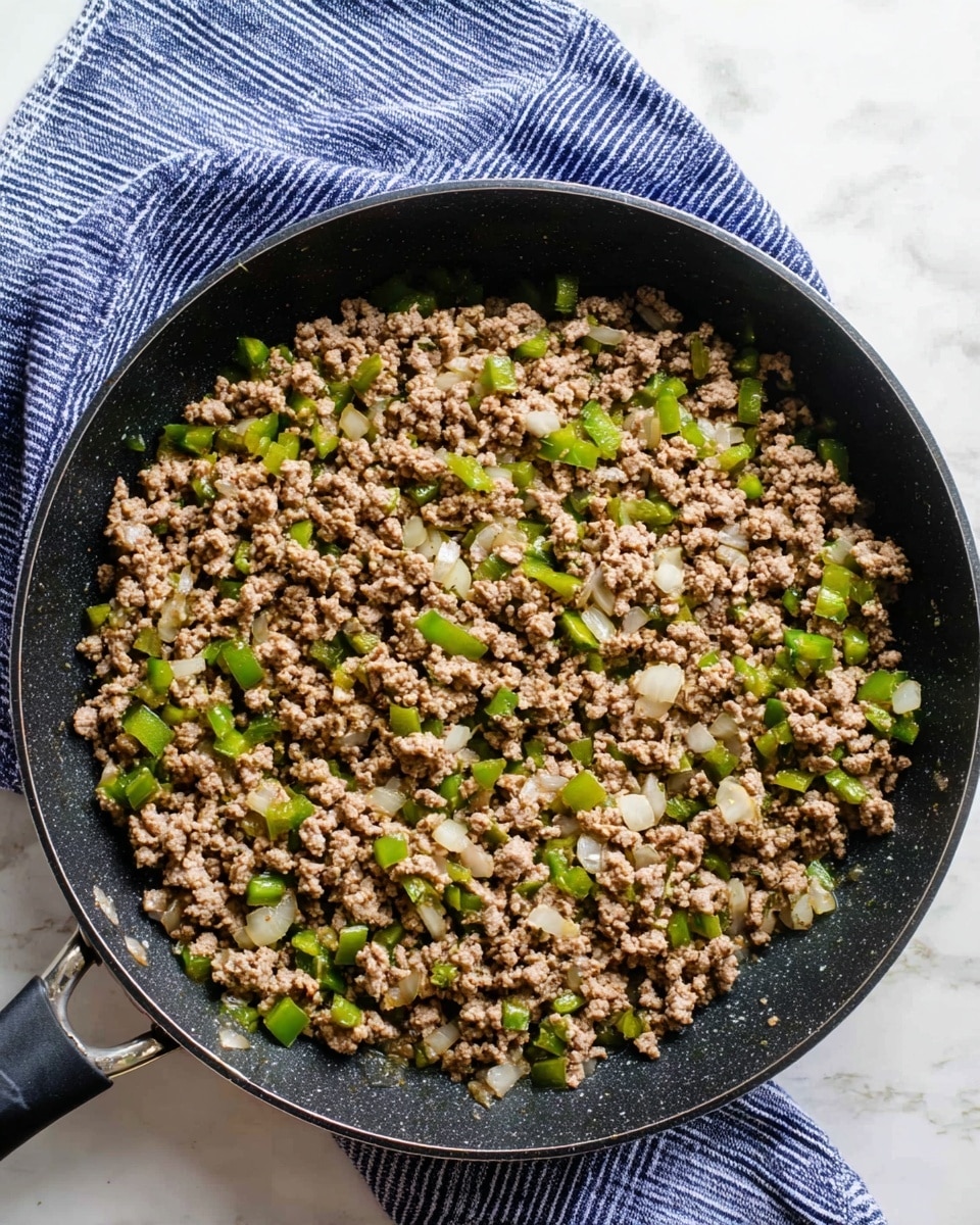 A black frying pan filled with cooked ground meat mixed with small pieces of green bell pepper and white onion, evenly spread across the pan's surface, showing a mix of light brown, green, and white colors with a slightly moist texture. The pan handle is visible on the left side, and a blue and white striped cloth is placed at the top edge on a white marbled surface. photo taken with an iphone --ar 4:5 --v 7