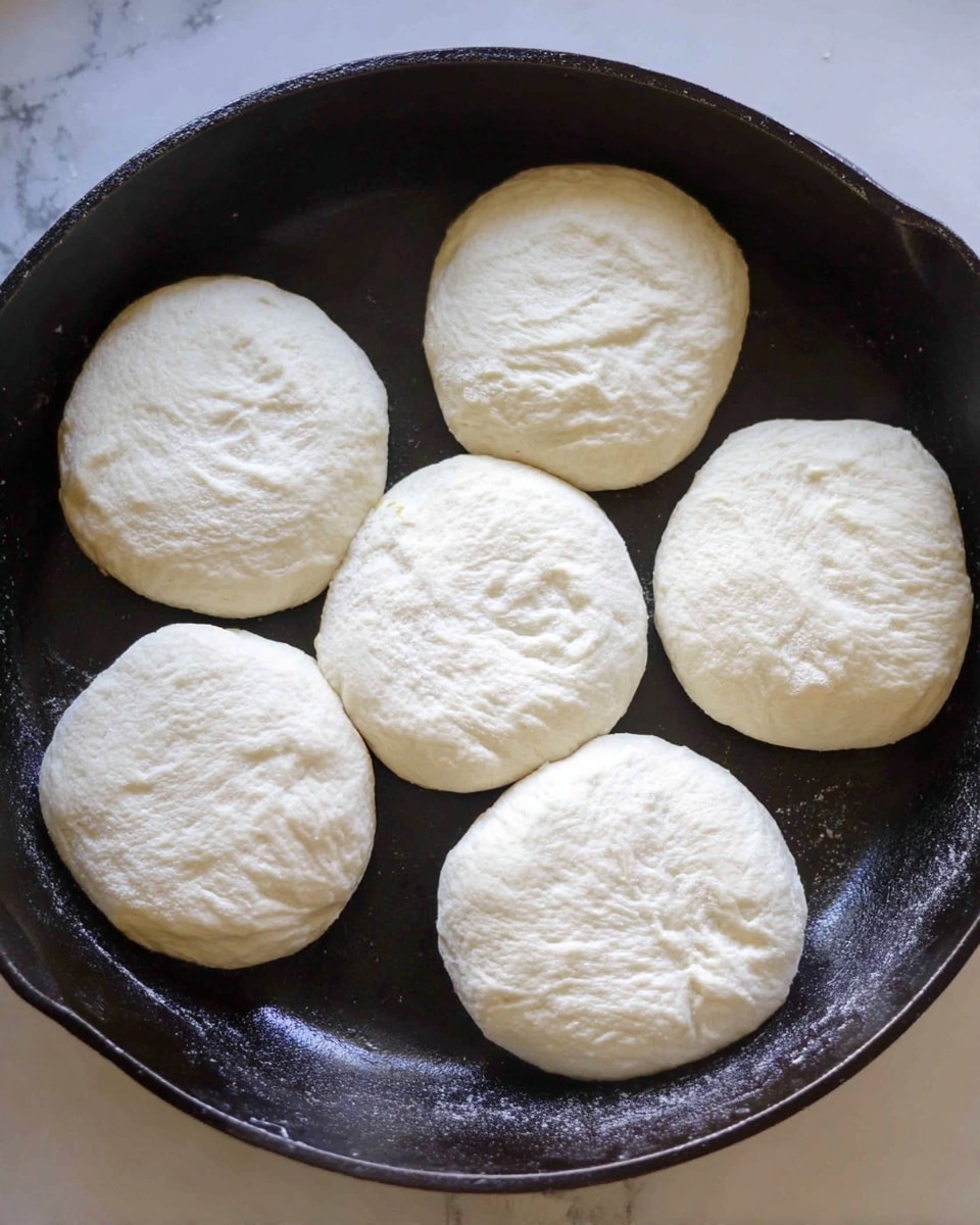 The image shows several toasted English muffins on a white marbled surface, some whole and some split open with a layer of dark red jam spread on the cut side. The toasted muffins have a light golden brown top with soft, white textured insides. A silver knife with some jam smeared on its blade lies near the muffins, with a small pile of jam next to it. In the background, a white plate holds more whole English muffins resting on a light blue and white striped cloth. The overall scene has a bright and soft lighting with a clean look photo taken with an iphone --ar 4:5 --v 7