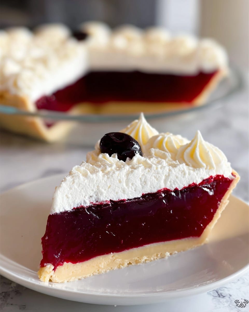 The image shows a slice of deep red jelly pie on a white plate resting on a white marbled surface. The pie has three layers: a thin pale yellow crust on the bottom, a thick smooth red jelly filling in the middle, and a thick layer of white whipped topping on top. The whipped topping is decorated with a swirl and a single dark berry placed in the center. In the background, the full pie with the same layers is visible out of focus. Photo taken with an iphone --ar 4:5 --v 7