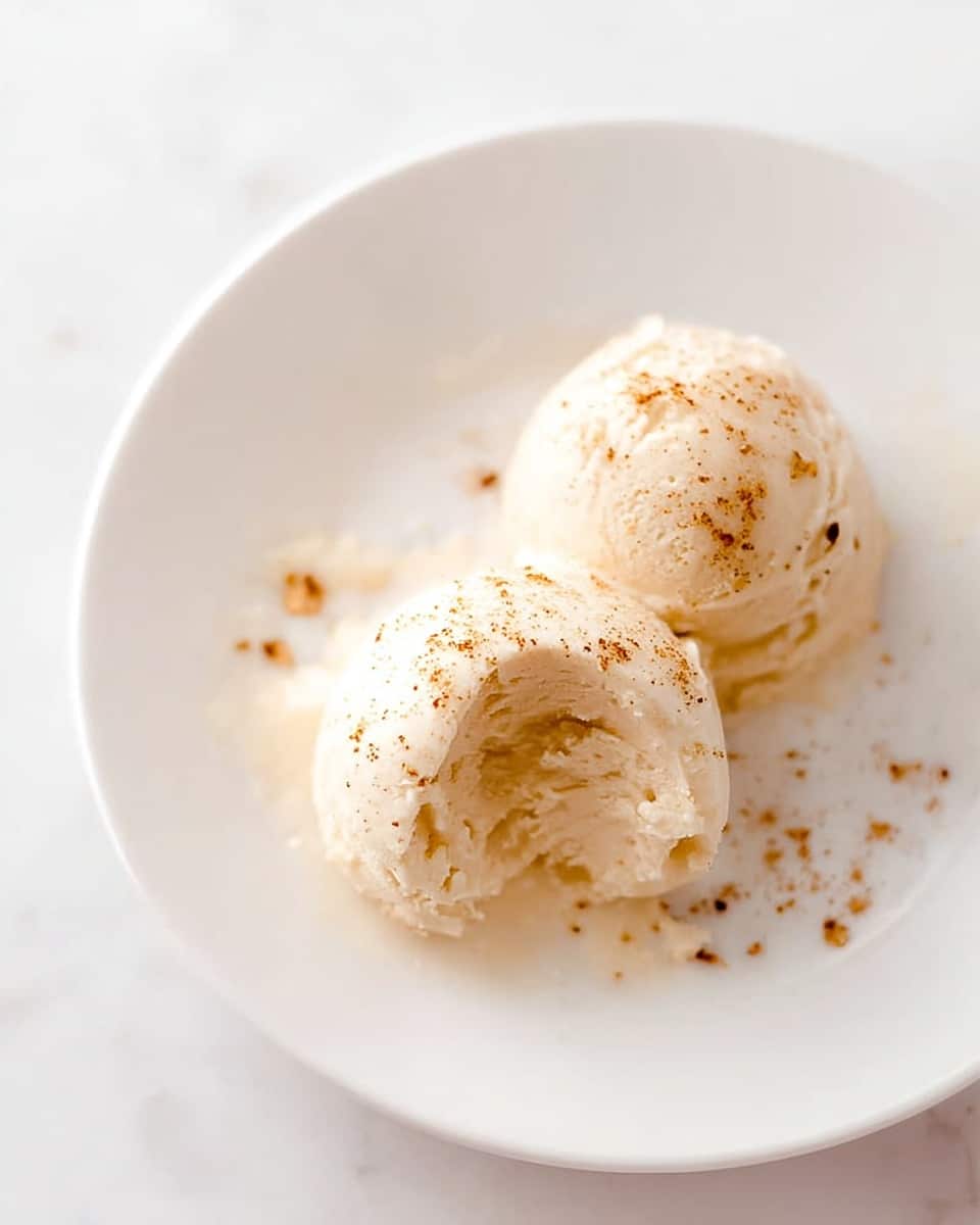 Two small round scoops of light beige ice cream sit on a white plate placed on a white marbled surface. The ice cream has a smooth, creamy texture with tiny brown specks sprinkled on top. One scoop is whole, while the other has a bite taken out of it, showing a soft, slightly textured inside. There are a few small crumbs and specks scattered lightly around the scoops on the plate. photo taken with an iphone --ar 4:5 --v 7