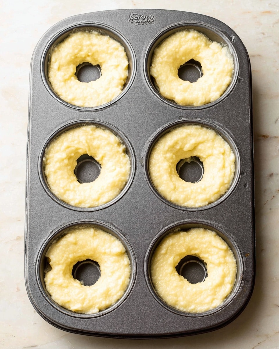 A dark gray metal donut baking tray with six circular molds filled with pale yellow batter that looks thick and lumpy. Each mold has a hole in the center, showing the tray's bottom. The batter fills each mold almost to the edge but is uneven on top, giving a rough texture. The tray is placed on a white marbled surface. photo taken with an iphone --ar 4:5 --v 7