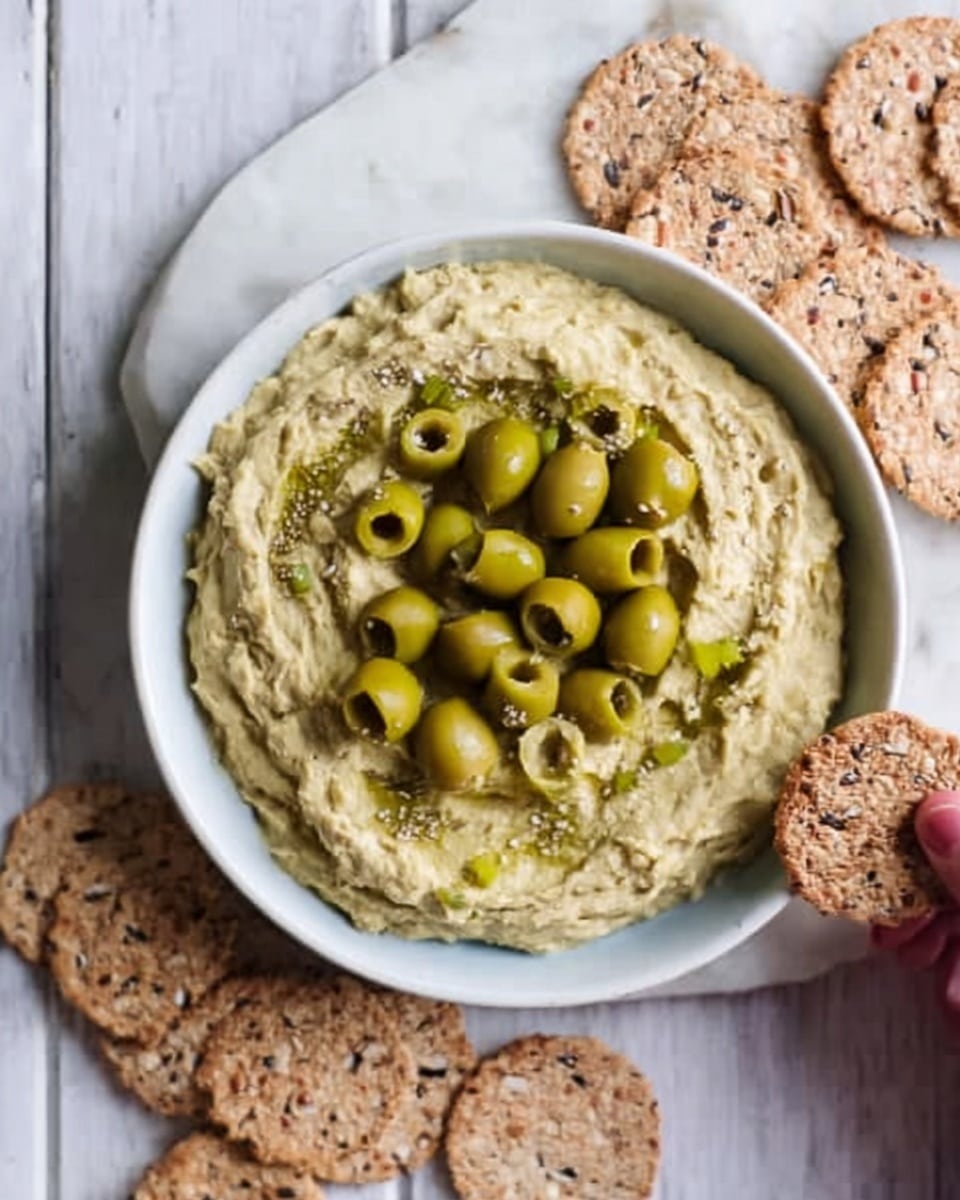 A white bowl filled with a creamy greenish-yellow dip topped with sliced green olives scattered evenly on the surface. The dip has a thick and slightly textured look. The bowl is placed on a white marbled surface, with a woman's hand reaching from the top right holding the edge of the bowl. Around the bowl, there are round, multigrain crackers with seeds visible on top, positioned mainly to the left and upper right side. Photo taken with an iphone --ar 4:5 --v 7