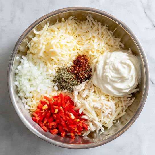In a large metal bowl, there is a mix of ingredients arranged in separate sections. At the base, shredded white cheese forms the main layer, topped with small piles of finely chopped white onion and bright red bell pepper pieces on one side. Next to them are thin strips of white cheese sprinkled with various dark spices. Small green chili pieces are also mixed with the spices. A large dollop of smooth, white sour cream sits near the front. Some shredded light-colored cooked chicken is placed over the shredded cheese as well. The bowl is set against a white marbled surface. Photo taken with an iphone --ar 4:5 --v 7
