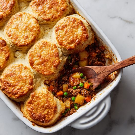 The image shows a white baking dish filled with a layered dish topped with thick, golden-brown biscuits. The biscuits have a fluffy texture with a crisp top layer and soft, pale beige side layers. Below the biscuits, the filling consists of a mixture of browned ground meat, diced orange carrots, yellow potatoes, and green peas, all coated in a rich, reddish-brown sauce. A wooden spoon is scooping one biscuit and some filling from the dish, highlighting the contrast between the soft biscuit top and the chunky, colorful filling beneath. The scene is set on a white marbled surface. photo taken with an iphone --ar 4:5 --v 7
