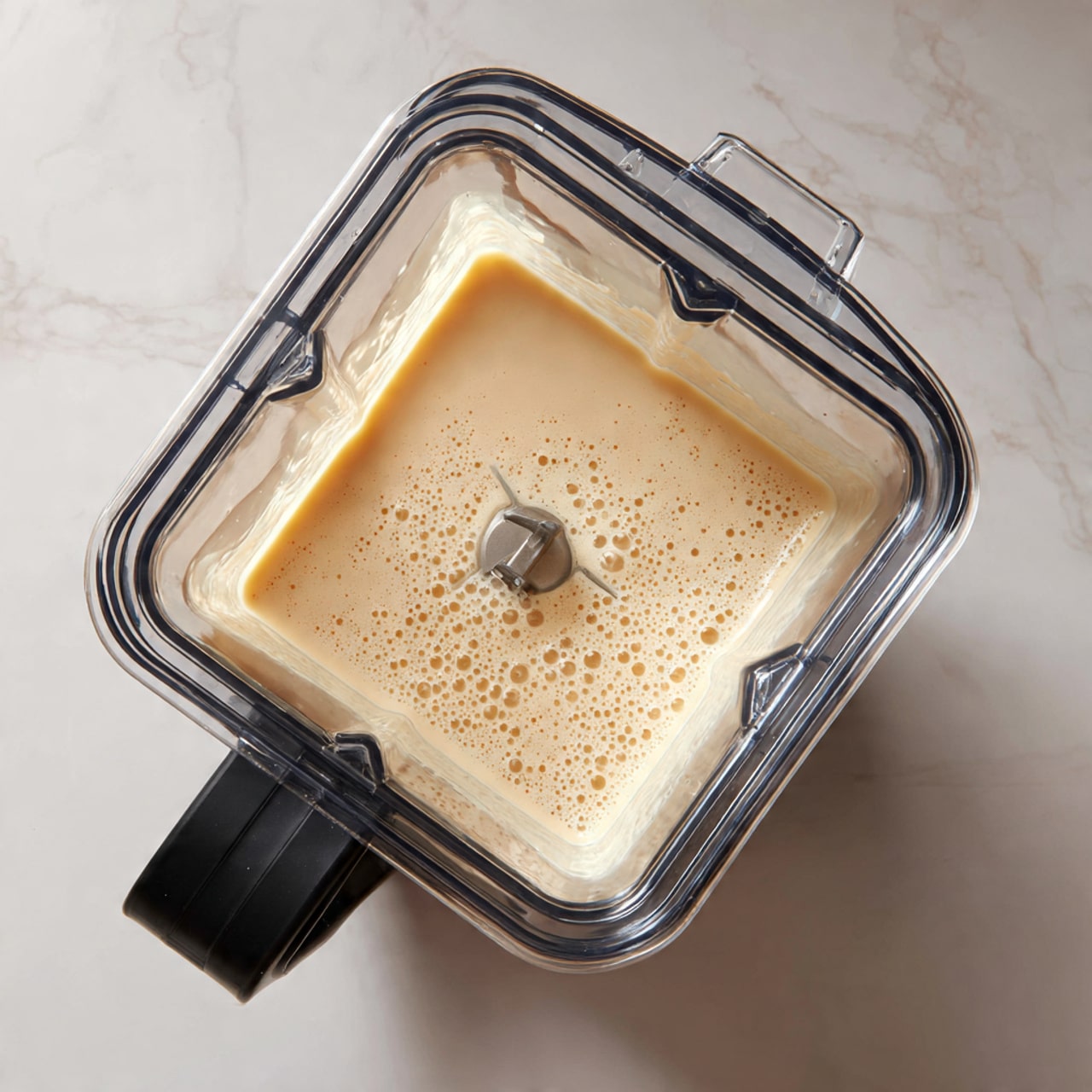 The image shows a square blender container with a black handle, filled with a light beige, smooth liquid with small bubbles on the surface, placed on a white marbled surface. The lid of the blender is off and lying nearby, showing the inside of the transparent cover. The blender blade is visible in the middle of the liquid. Photo taken with an iphone --ar 4:5 --v 7
