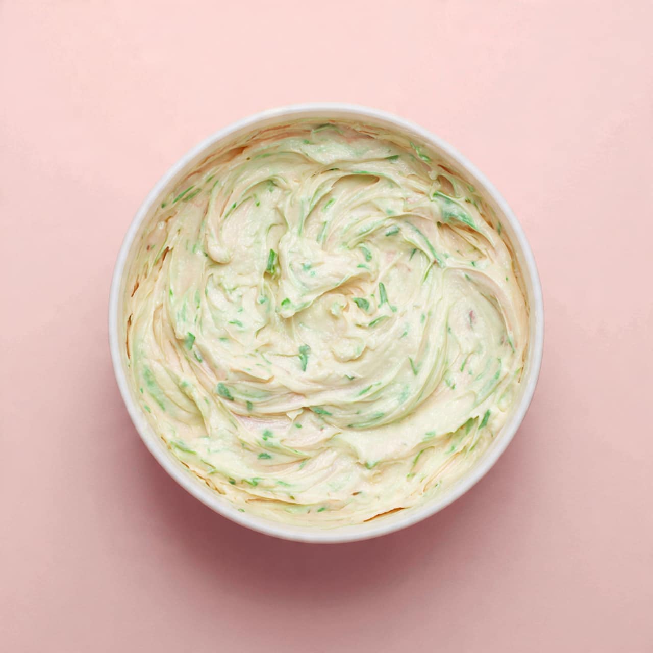 A white bowl filled with a creamy white mixture that has visible green herb pieces mixed throughout. The mixture has a smooth, soft texture with some gentle swirls on the surface, showing it has been stirred or whipped. The bowl is viewed from above, sitting on a soft pink background. photo taken with an iphone --ar 4:5 --v 7
