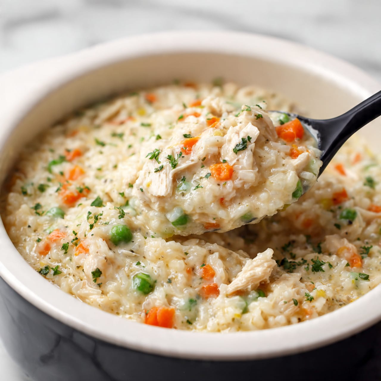 A white plate holds a creamy risotto dish with three main layers visible: at the base, small, soft risotto grains in a light beige color mixed evenly throughout; scattered on top are small, vibrant orange pieces of carrot and bright green peas adding color contrast; creamy shredded white meat pieces are layered within the risotto, all mixed in with fresh green parsley sprinkled over the top for a finishing touch. The risotto is lightly topped with finely grated white cheese, giving a delicate texture on the surface. There is a shiny silver spoon resting on the left side of the plate, partially sunk into the risotto. The plate sits on a white marbled surface. photo taken with an iphone --ar 4:5 --v 7