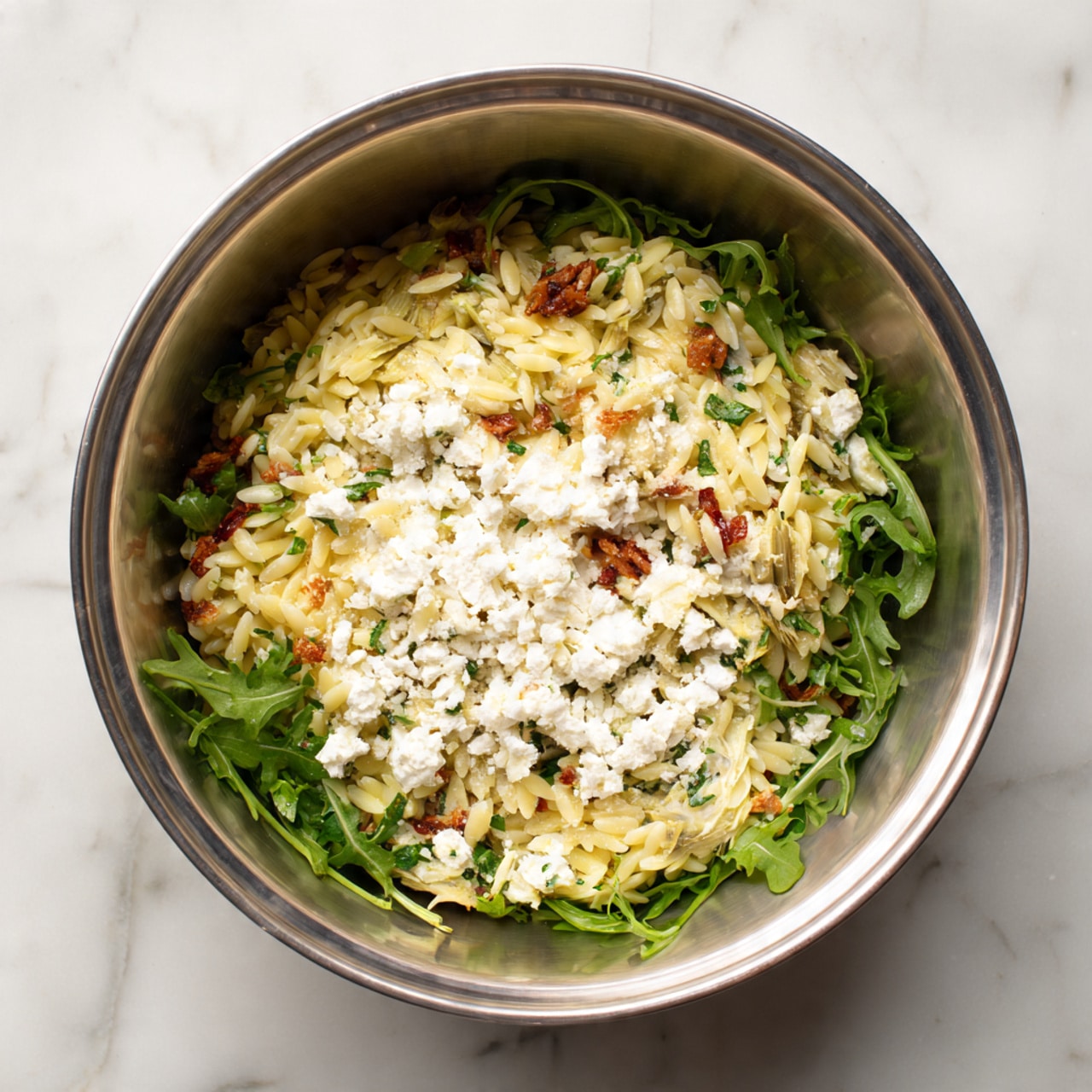 A large wooden bowl filled with orzo pasta salad that has light yellow orzo as the base layer, mixed with green arugula leaves scattered throughout, white pieces of crumbled cheese, pale artichoke hearts, and bright red sun-dried tomato strips evenly spread in the dish. Two wooden spoons rest in the bowl, slightly submerged in the salad, with some orzo and sun-dried tomatoes on them. The scene is set on a white marbled texture surface photo taken with an iphone --ar 4:5 --v 7