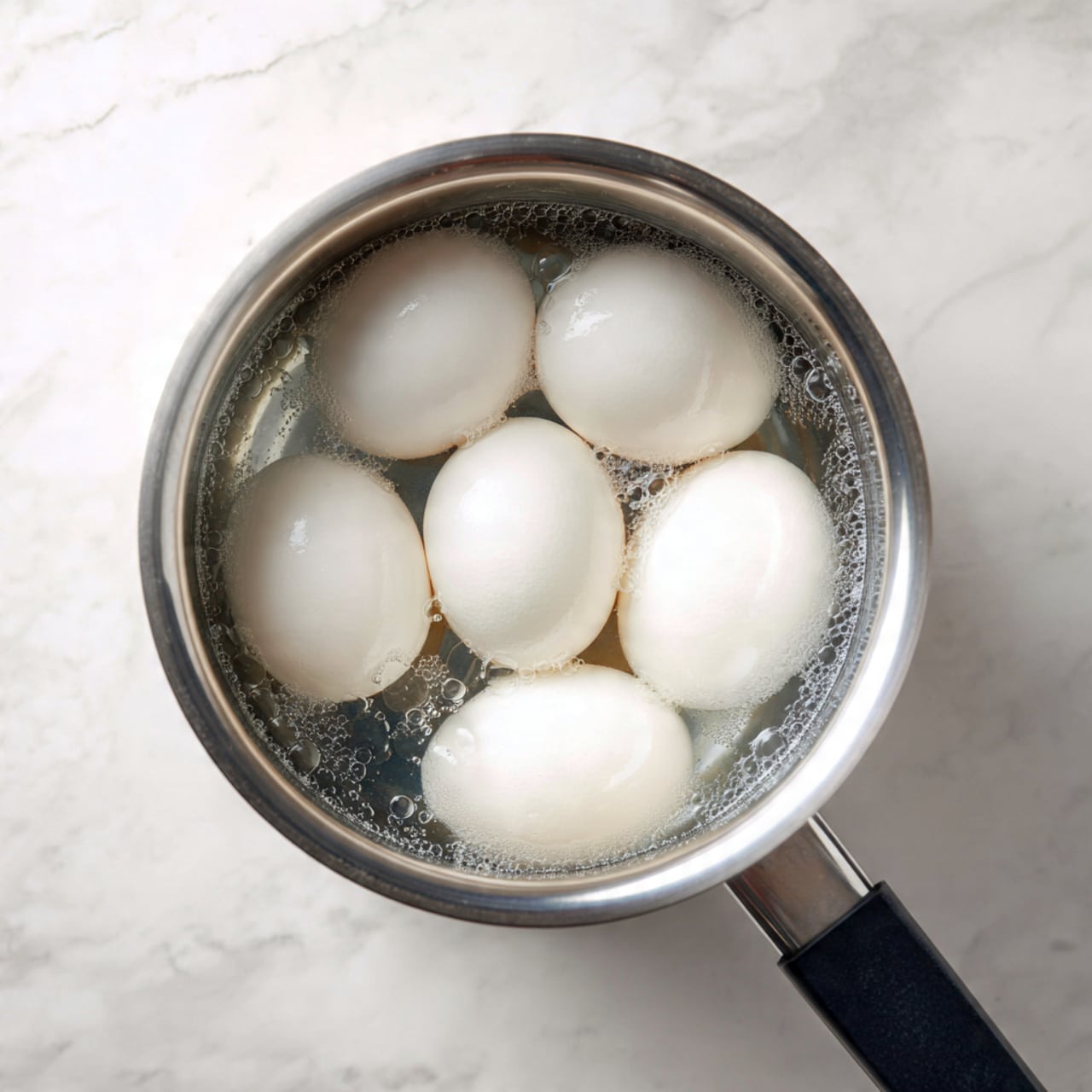 A top view of a metal pot filled with water and six white eggs fully submerged inside. The pot has a shiny silver inside texture and a black handle on the right side. The pot is placed on a white marbled surface with soft natural light illuminating the scene, creating gentle reflections on the eggs and the water surface. photo taken with an iphone --ar 4:5 --v 7