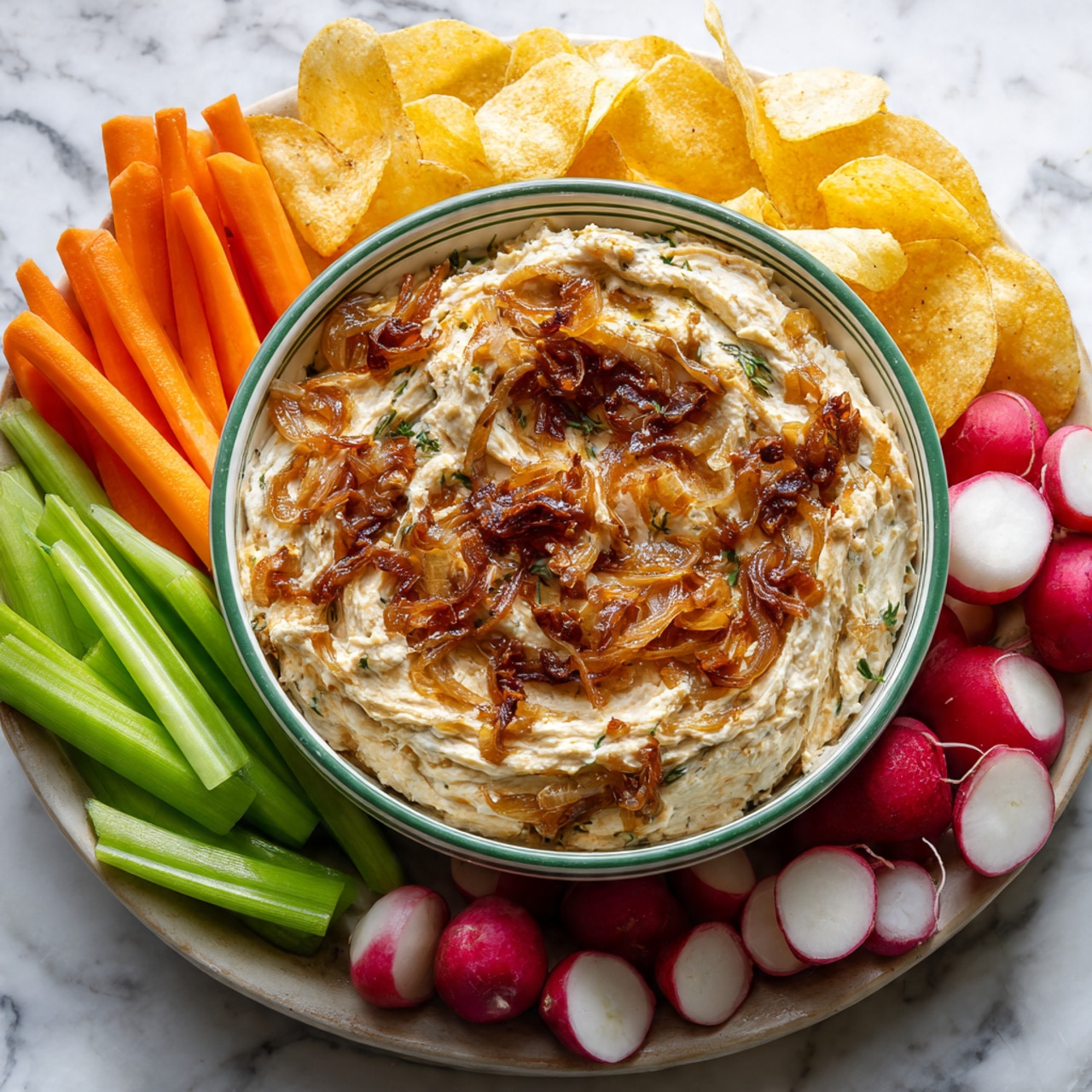 The image shows a round white bowl with green stripes filled with a creamy dip that has mixed bits of caramelized onions, giving it a swirled texture. The dip is thick and light beige with reddish-brown onion pieces spread on top. Around the bowl, there are light yellow potato chips, bright orange carrot sticks, green celery sticks, and fresh red radishes with white interiors arranged on a white marbled surface. photo taken with an iphone --ar 4:5 --v 7