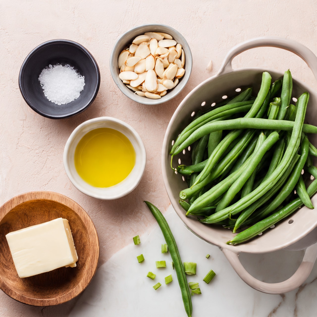 The image shows fresh green beans inside a light gray colander on the right side, with some green bean ends placed on the white marbled surface nearby. To the left are four small bowls arranged horizontally: a black bowl with white salt, a light gray bowl filled with sliced almonds, a white bowl with olive oil, and a small wooden bowl holding a chunk of butter. The background is a smooth pink surface. photo taken with an iphone --ar 4:5 --v 7