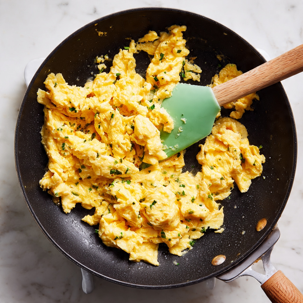 A close-up of a black frying pan with soft, yellow scrambled eggs cooking inside. The eggs have a slightly shiny, smooth texture with some small curds and are gathered mostly in the center of the pan. A green spatula with a light wooden handle is held from the side, lifting part of the eggs. The pan rests on a white marbled surface with a small part of a white stovetop visible at the top right corner. Photo taken with an iphone --ar 4:5 --v 7