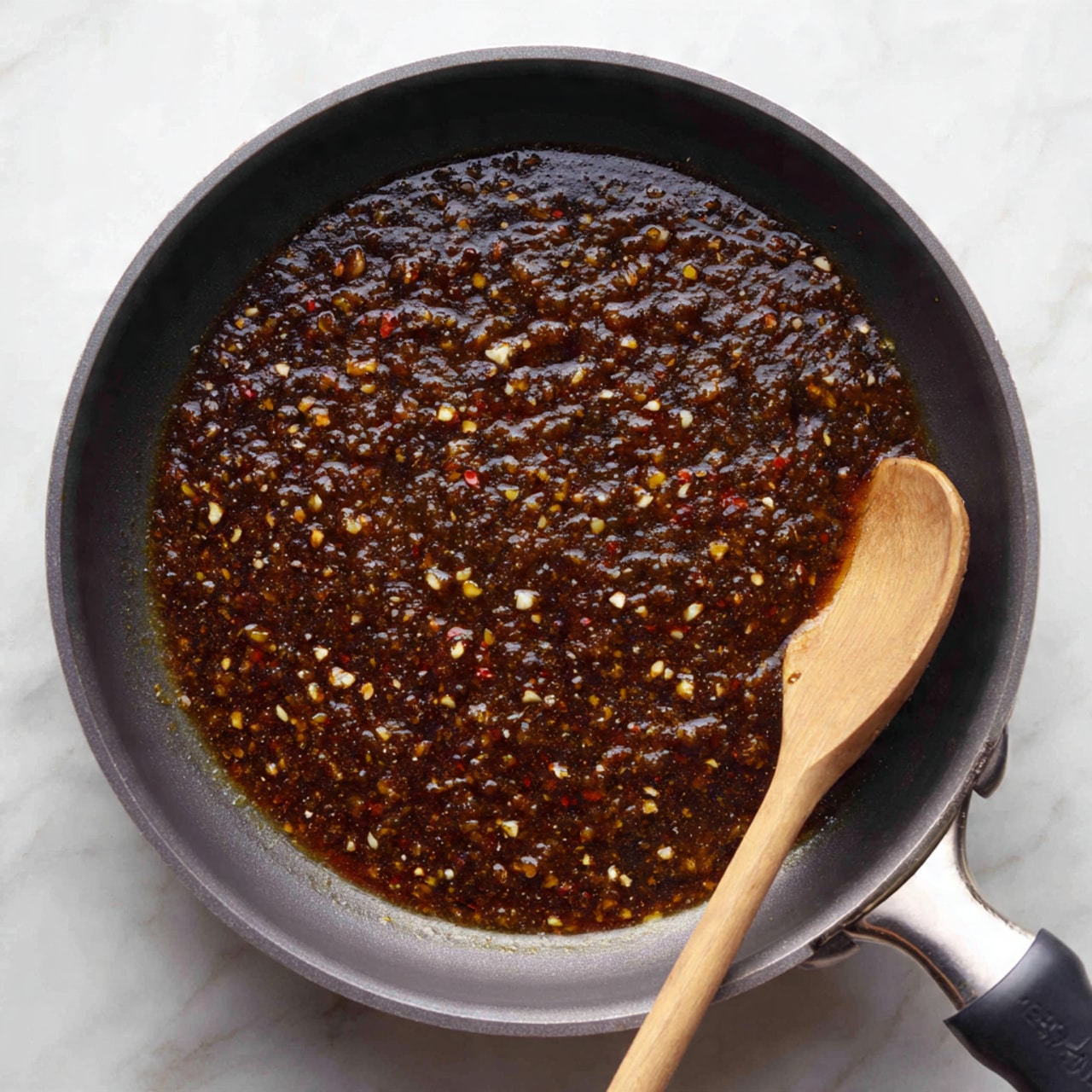 A close-up view of a black frying pan filled with a thick, dark brown sauce mixed with small white and yellow bits, likely garlic and spices. The sauce has a glossy texture and looks stirred, with a light-colored wooden spoon resting inside the pan on the right side. The pan itself has a light gray rim and a black handle on the right. The background is a white marbled surface. Photo taken with an iphone --ar 4:5 --v 7