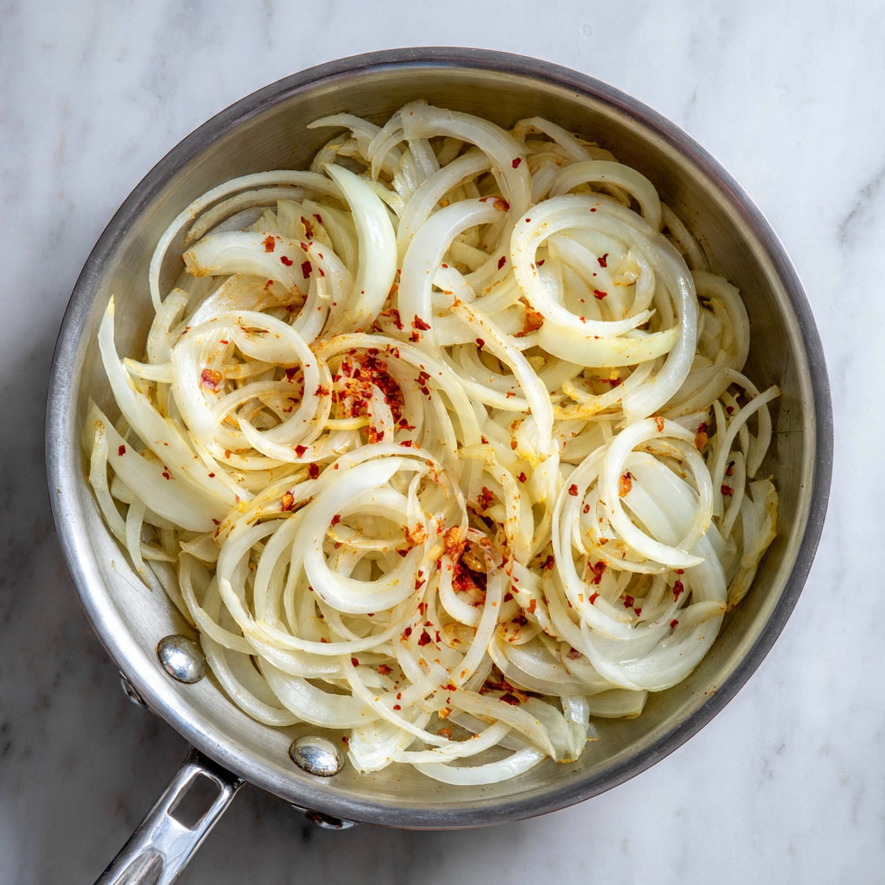 A silver pan filled with long, thin, spiral-shaped white onion slices arranged loosely on top of each other. The onions have a light texture and are sprinkled with small patches of red seasoning in the center. The pan sits on a white marbled surface. photo taken with an iphone --ar 4:5 --v 7