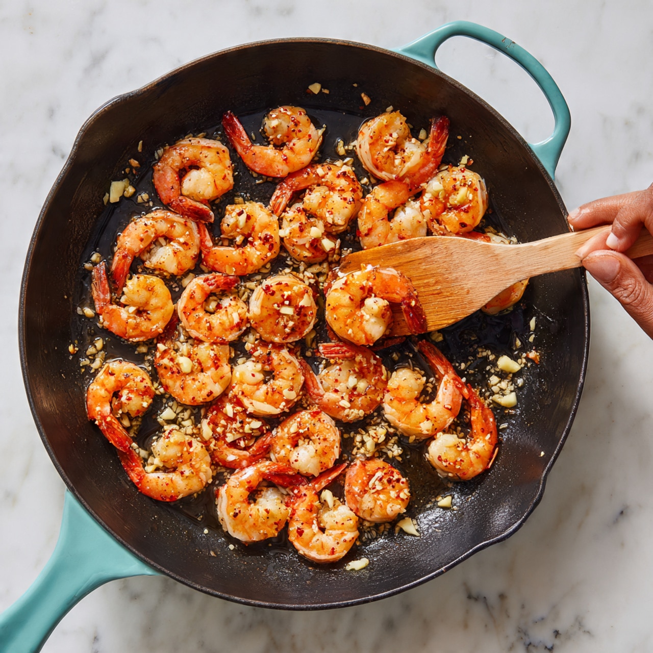 A black cast iron pan sits on a white marbled surface, filled with about twenty shrimp cooking. The shrimp are arranged in a loose circular pattern, turning pinkish-orange with some red spice specks on their bodies. Small pieces of chopped garlic are scattered among the shrimp, showing a light golden color. A wooden spatula held by a woman's hand is lifting a shrimp on the right side of the pan. The pan handles are light teal, contrasting with the dark pan and bright shrimp. Photo taken with an iphone --ar 4:5 --v 7