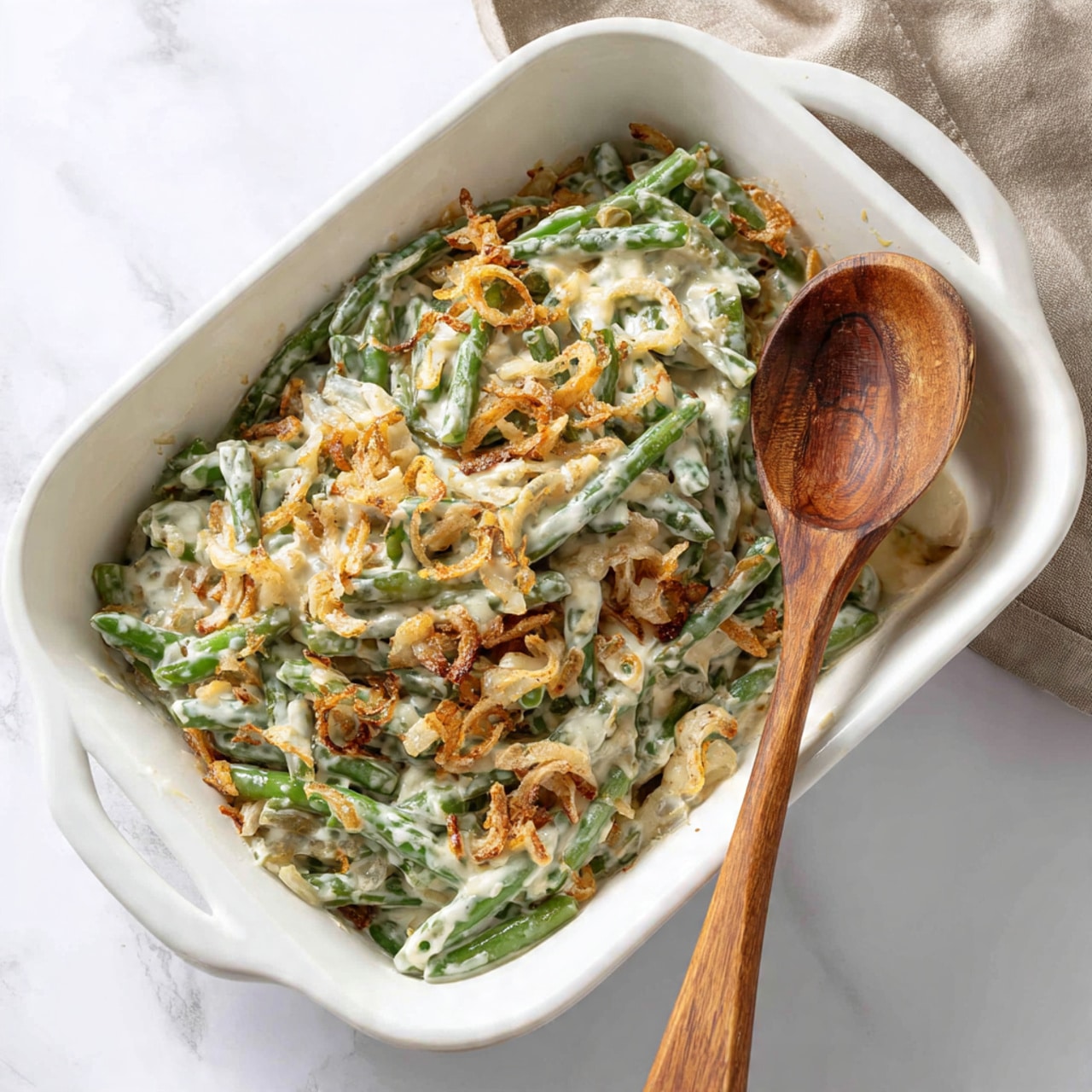 A white rectangular baking dish filled with a creamy green bean casserole. The casserole has one visible layer, with green beans mixed with a thick white sauce that has a smooth texture, speckled with small bits of light brown fried onions scattered on top and inside. A wooden spoon rests inside the dish on the right side, partially covered by the creamy mixture. The dish is placed on a white marbled surface. Photo taken with an iphone --ar 4:5 --v 7
