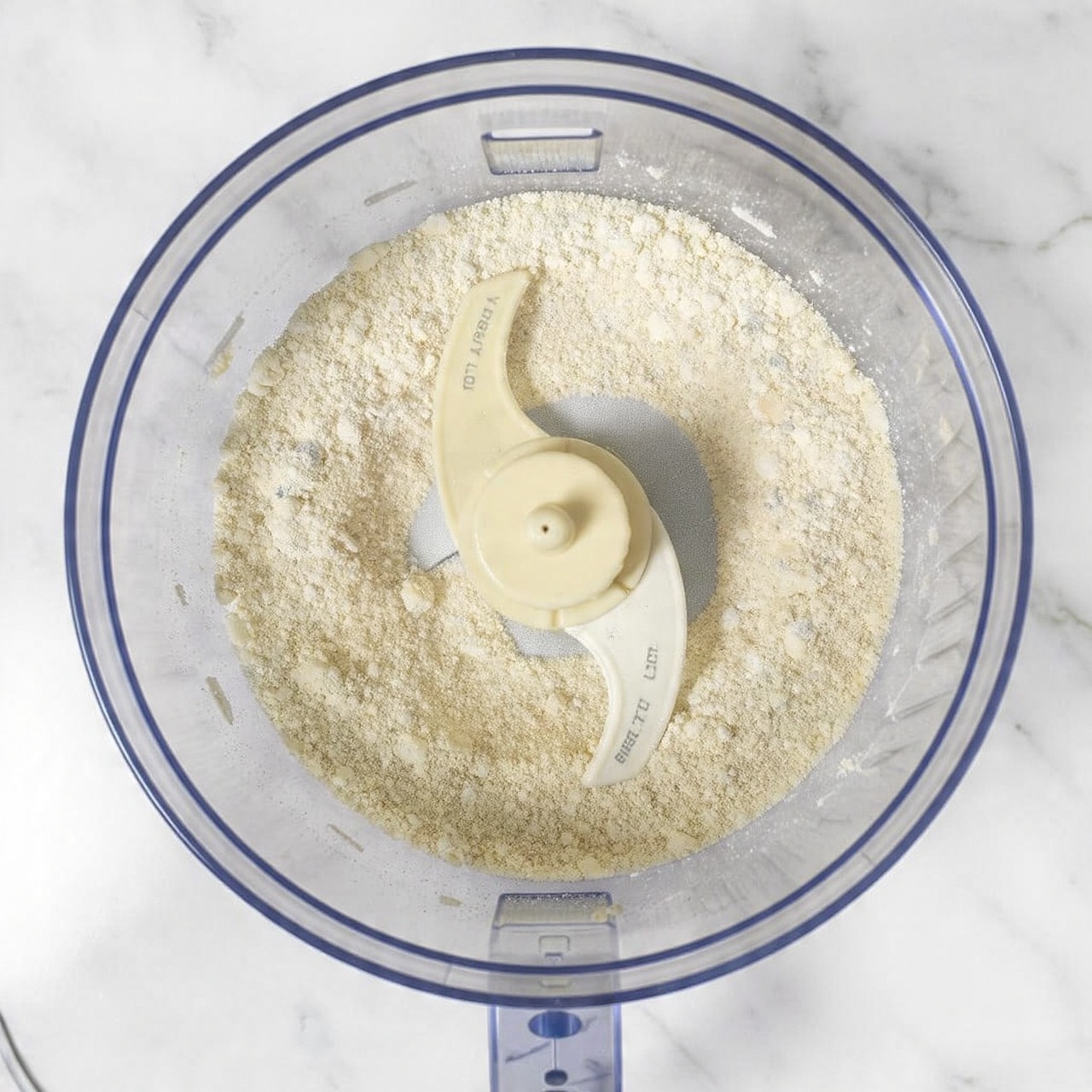 Top view of a clear food processor bowl filled with a mixed dry flour and crumbly white fat mixture, showing a rough, grainy texture evenly spread inside. The center of the bowl has a beige plastic blade guard with some text on it. The bowl sits on a white marbled surface. Photo taken with an iphone --ar 4:5 --v 7