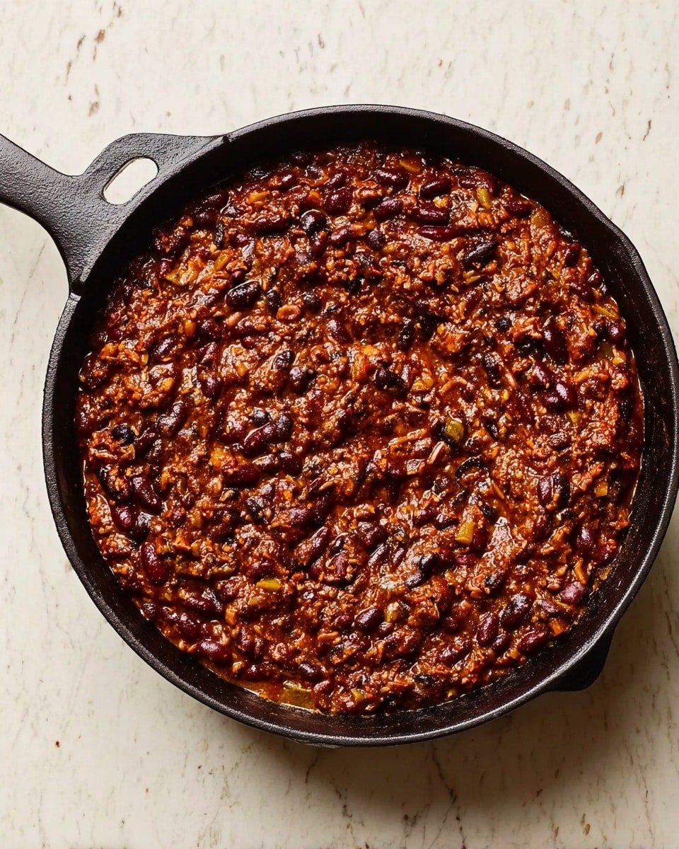 A black cast iron skillet filled with a thick, dark brown mixture that looks like a spicy black bean stew, with visible black beans, small bits of green herbs, and a rich reddish sauce. The stew is slightly swirled on the surface, showing a dense and hearty texture. The skillet is placed on a white marbled surface background. photo taken with an iphone --ar 4:5 --v 7