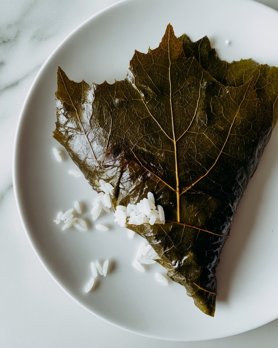 The image shows three dark green stuffed grape leaves placed side by side on a white rectangular plate. Next to the grape leaves are two bright yellow lemon wedges. Around the plate, fresh green mint leaves are scattered on a white marbled surface. In the background are blurry green herbs and a white bowl containing red cherry tomato halves. Photo taken with an iphone --ar 4:5 --v 7