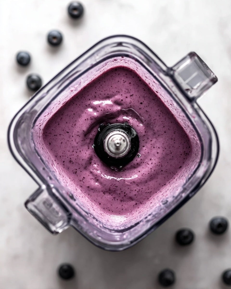 A close-up image shows a thick purple smoothie being poured from a blender into a clear glass, filling it to the top. The smoothie has a creamy texture with tiny dark specks throughout. Behind, another glass filled with the same smoothie is slightly out of focus. The white marbled surface holds scattered fresh blueberries and small green spinach leaves. In the corner, a white bowl with seeds adds detail. The background is bright with soft shadows, and a woman's hand holds the blender while pouring. Photo taken with an iphone --ar 4:5 --v 7
