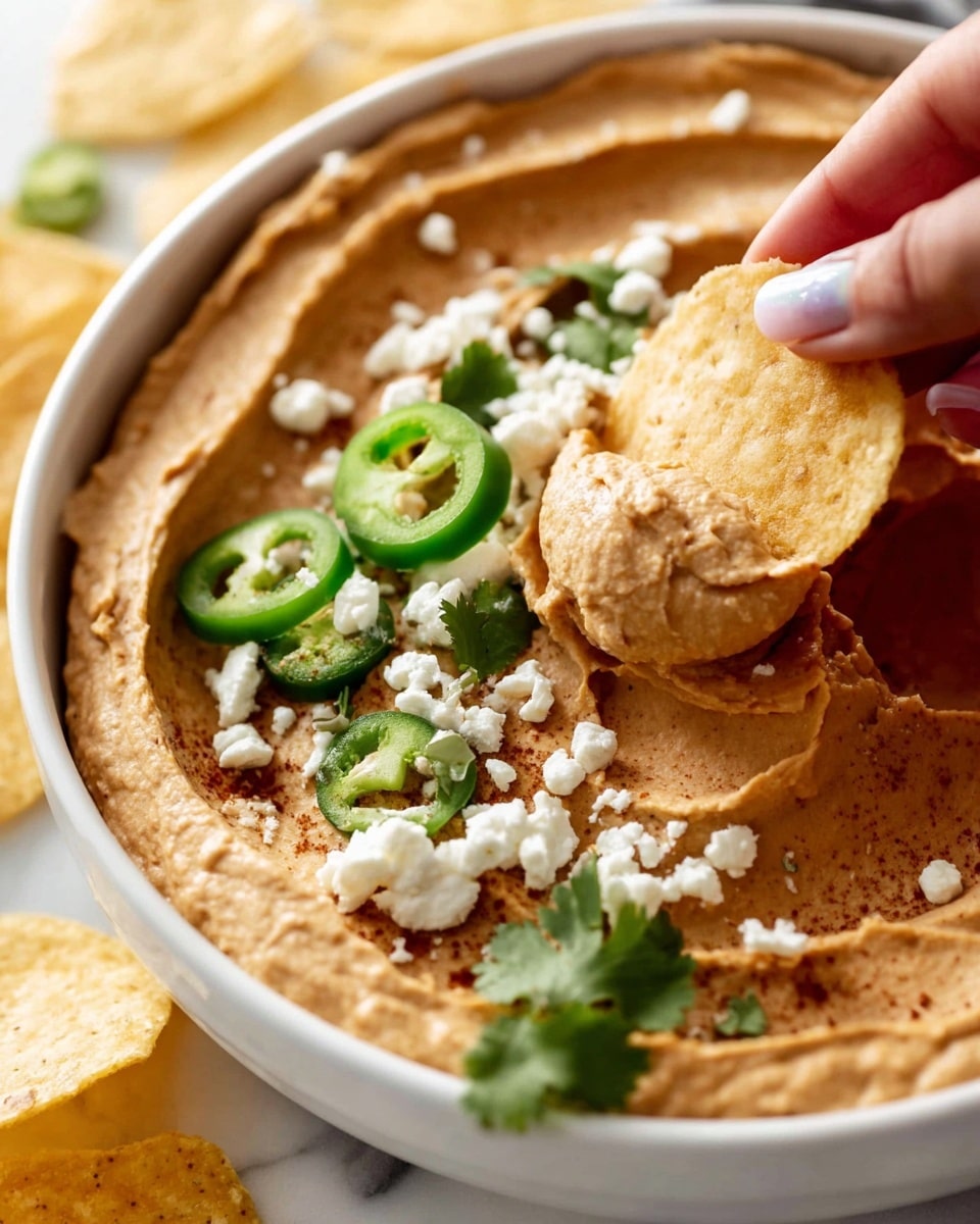 A close-up of a white bowl filled with smooth, light brown creamy dip spread evenly with gentle swirls, topped with small white crumbly cheese pieces, thin green jalapeño slices, and fresh green cilantro leaves scattered on top. A woman's hand holding a round, lightly toasted beige chip is dipping into the creamy layer at the right side of the bowl. The background shows additional chips on a white marbled surface. photo taken with an iphone --ar 4:5 --v 7