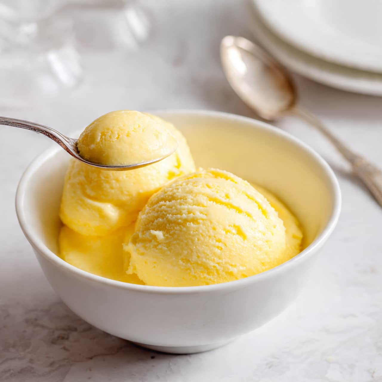 A large white plate filled with a smooth yellow mango mousse that takes up most of the center. The top edge is decorated with bright orange mango slices arranged in a curved line, interspersed with small dollops of white whipped cream. A silver spoon rests in a scooped-out section on the left side of the mousse. Around the plate, there are smaller white dishes, one holding more mango slices and another with additional mousse and a spoon, all placed on a white marbled surface. A dark blue cloth is partially visible in the bottom right corner. Photo taken with an iphone --ar 4:5 --v 7