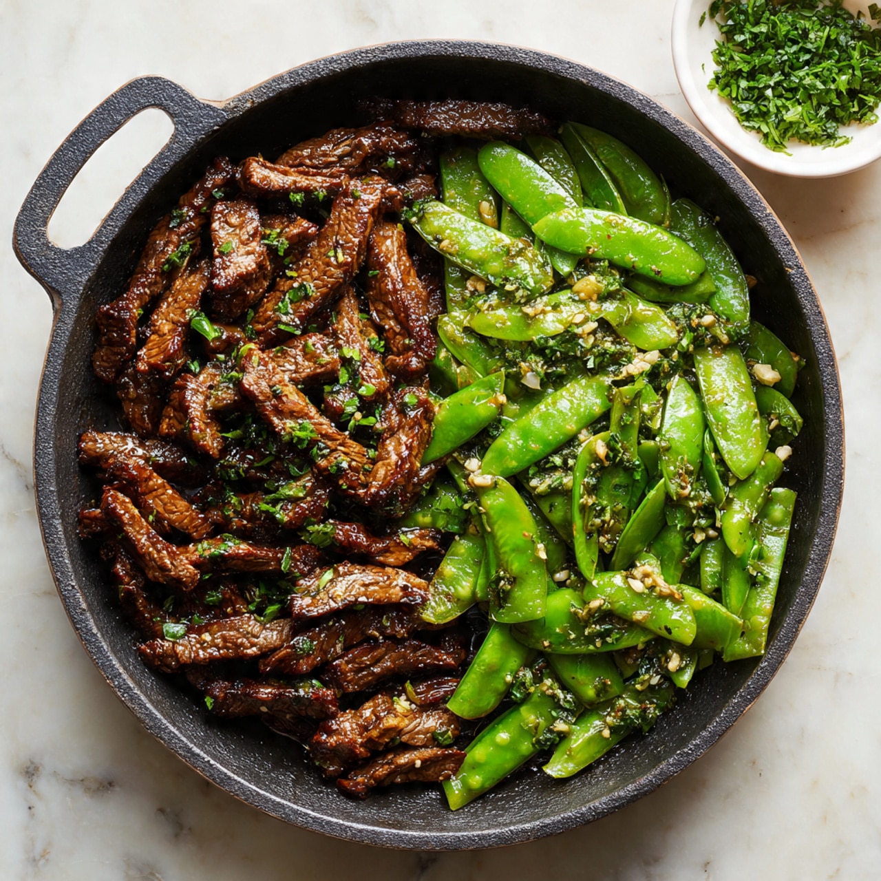 A black cast iron pan filled with two layers side by side: on the left, a layer of dark brown cooked sliced beef with a slightly shiny texture, sprinkled with small chopped green herbs; on the right, bright green snow peas with a slightly glossy surface mixed with small white bits, also topped with green herb pieces. In the upper right corner, a white bowl holds more chopped green herbs. The pan sits on a white marbled surface with a soft natural light. photo taken with an iphone --ar 4:5 --v 7