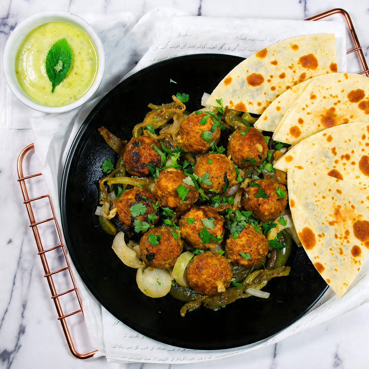 The image shows a white plate filled with eight dark brown grilled meatballs placed on a bed of cooked onions and green chili slices. The meatballs have a slightly charred texture with small bits of green herbs like cilantro sprinkled over them. The cooked onions beneath are soft, caramelized with golden and light brown colors. Some green coriander leaves are scattered on the plate, adding fresh green spots. In the background, blurred, there is a small bowl of white sauce with a mint leaf on top and pieces of flatbread resting behind the plate, all set on a white marbled surface. photo taken with an iphone --ar 4:5 --v 7