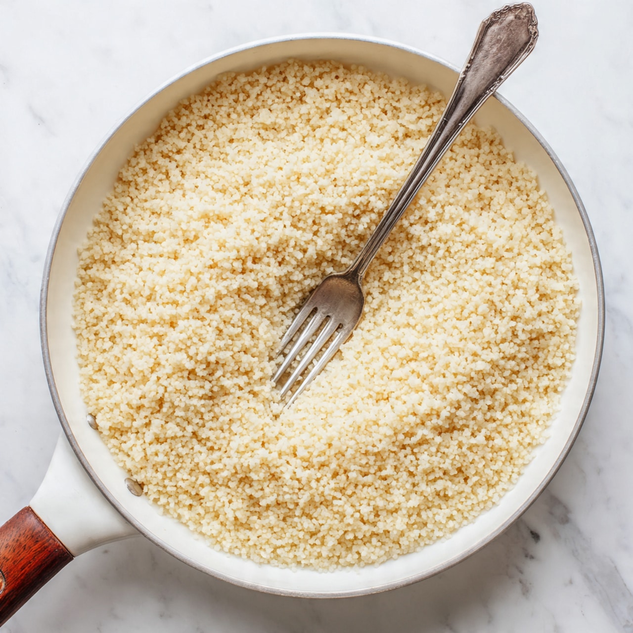 A close-up top view of a white pot with a wooden handle, filled with fluffy, light yellow couscous grains, showing a soft, grainy texture all over. There is a silver spoon with a curved handle placed inside the pot on the right side, partially sunk into the couscous. The pot is sitting on a white marbled surface with subtle grey veins. photo taken with an iphone --ar 4:5 --v 7