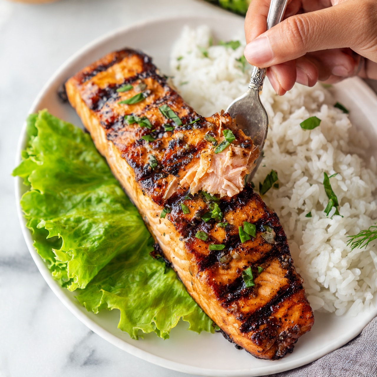 A white plate on a white marbled surface holds a grilled salmon fillet with dark brown grill marks and small green herb pieces on top. The salmon is pink inside, with a small piece being lifted by a silver fork held by a woman's hand. To the right of the salmon is a pile of white rice with a fluffy texture. Under the salmon, there are a few bright green lettuce leaves. The image is brightly lit, showing clear details of the textures and colors. photo taken with an iphone --ar 4:5 --v 7